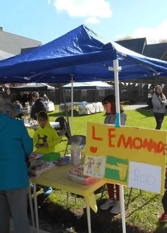 Community yard sale booth with a blue canopy, a yellow sign reading 'Lemonade,' and various items on the table in an outdoor residential neighborhood with houses and people, some shopping and some selling.