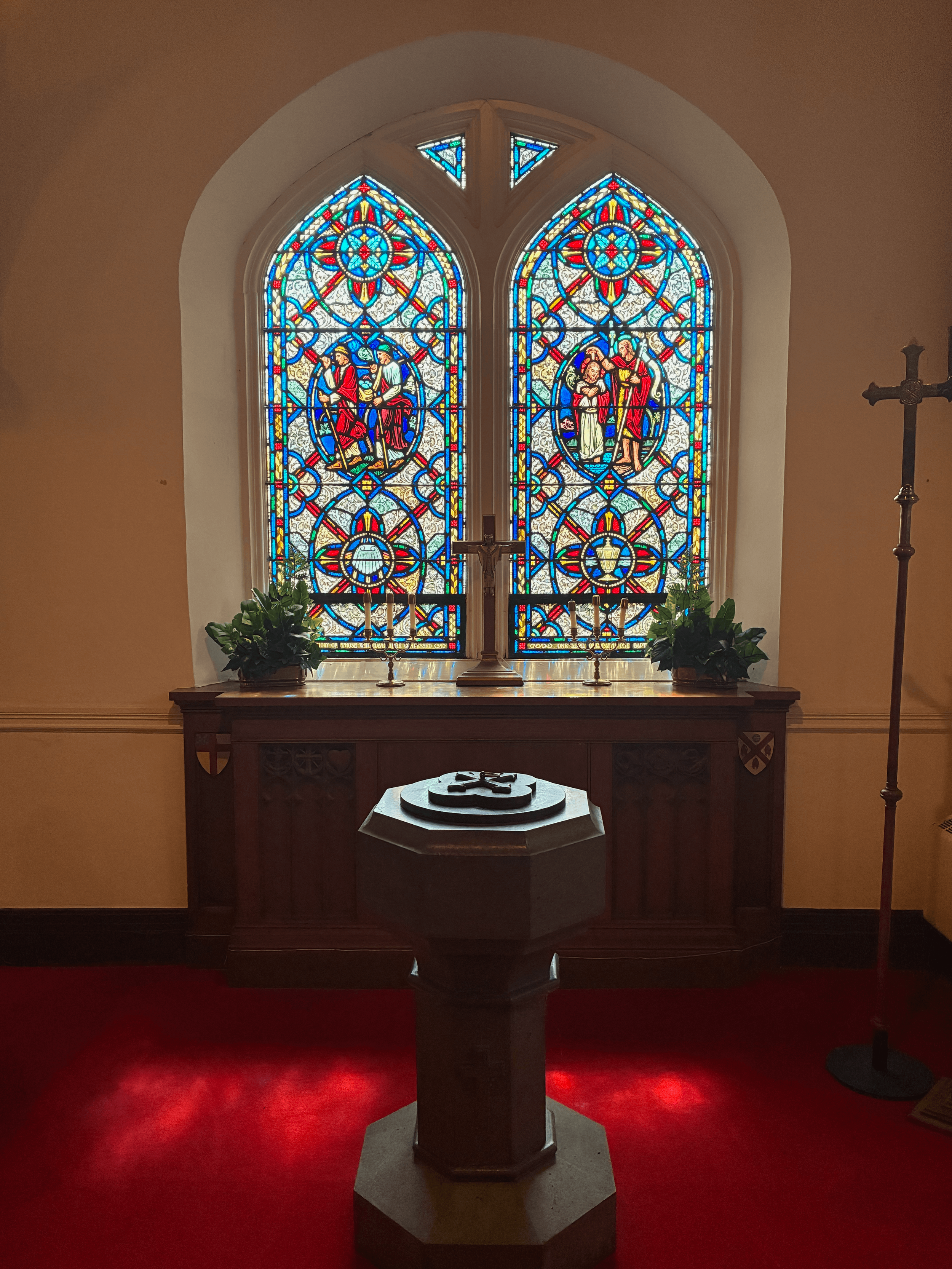 Inside a church, a stained glass window depicting scenes from the Bible with vibrant colors, a wooden altar with candles and potted plants, a cross on the right, and a bishop's chair on the left, all illuminated by sunlight.