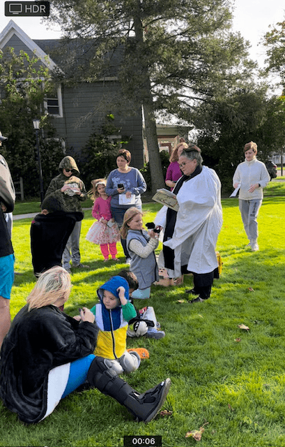A group of children and adults gathered outdoors on a grassy area during daytime, with some children sitting on the grass and some adults standing, one dressed in a white coat, possibly participating in an event or ceremony.