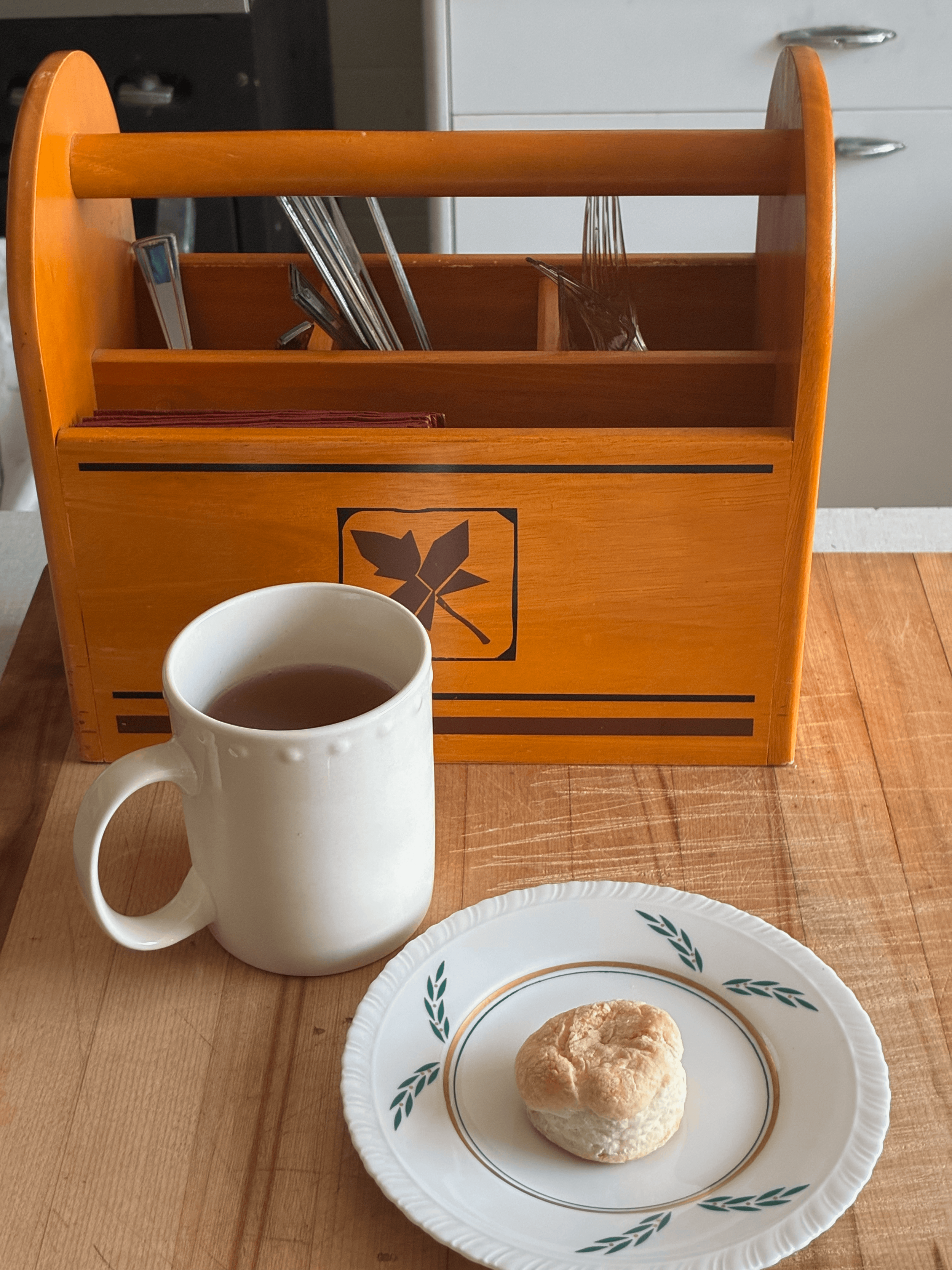 A white mug filled with coffee sitting on a light wooden table, with a white plate with green and gold leaf design holding a biscuit in front of a wooden dish organizer containing silverware and napkins.