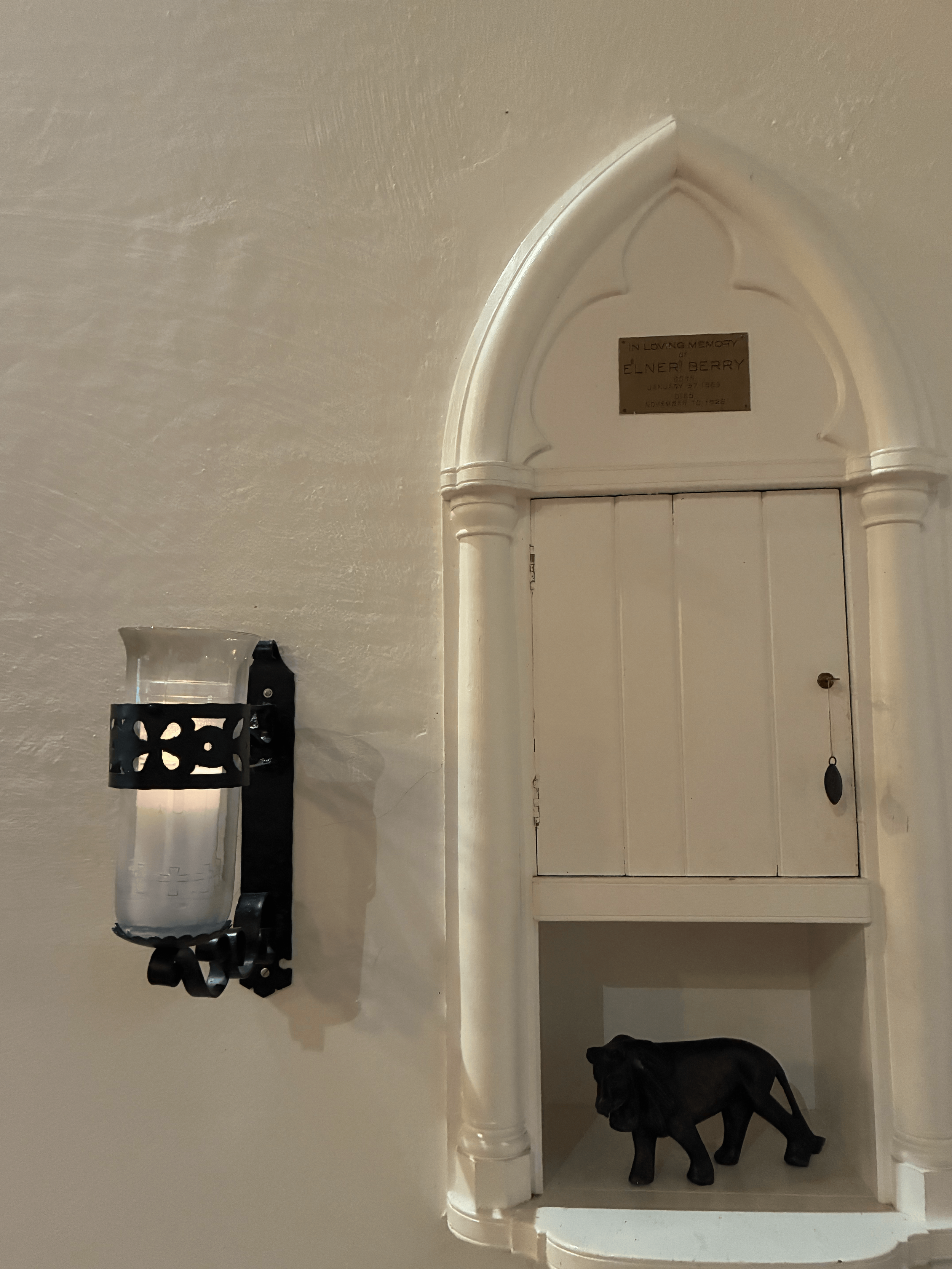 White wooden cabinet with a plaque reading 'In Loving Memory of Elner Berry January 27, 1883 to November 14, 1925'. A black metal wall sconce holds a glass candle holder, and a black dog is standing underneath the cabinet.