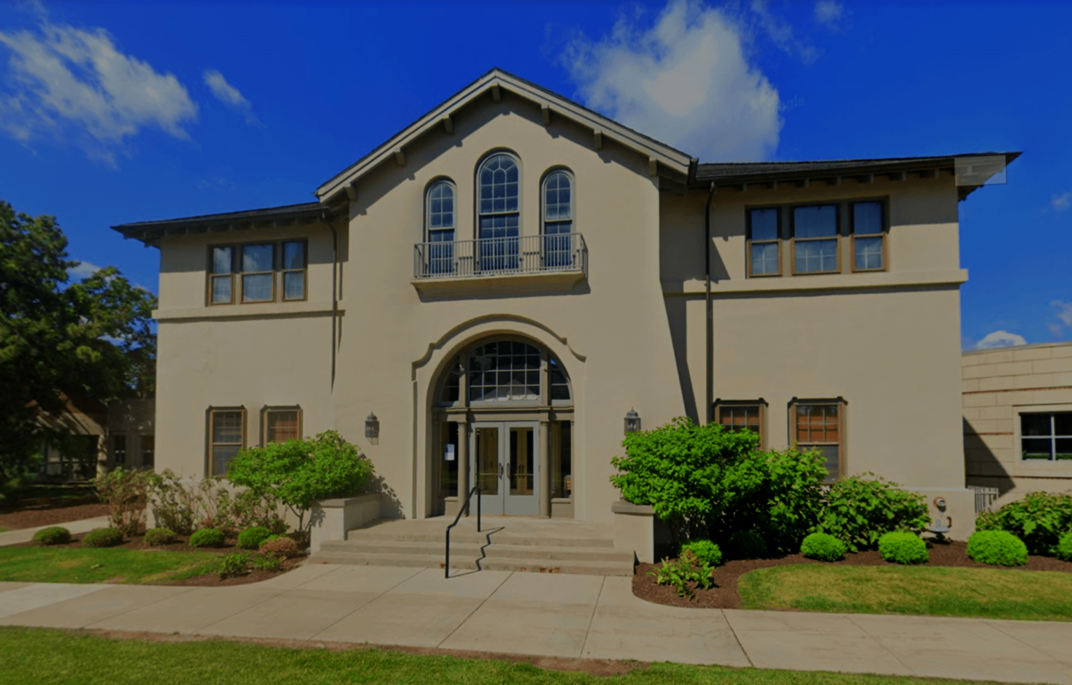 Front view of a beige, two-story house with arched windows, a small balcony, and a landscaped lawn with bushes and trees under a clear blue sky.