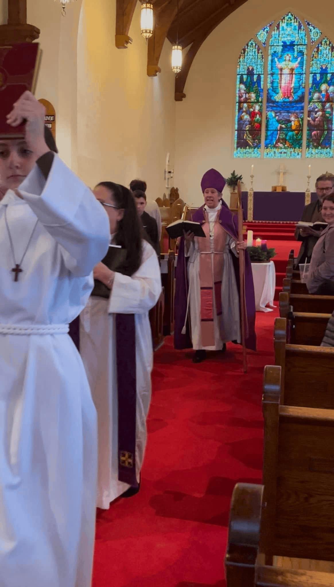 A religious ceremony inside a church with stained glass windows, a priest or bishop wearing purple robes, and several people in white robes participating in the service.