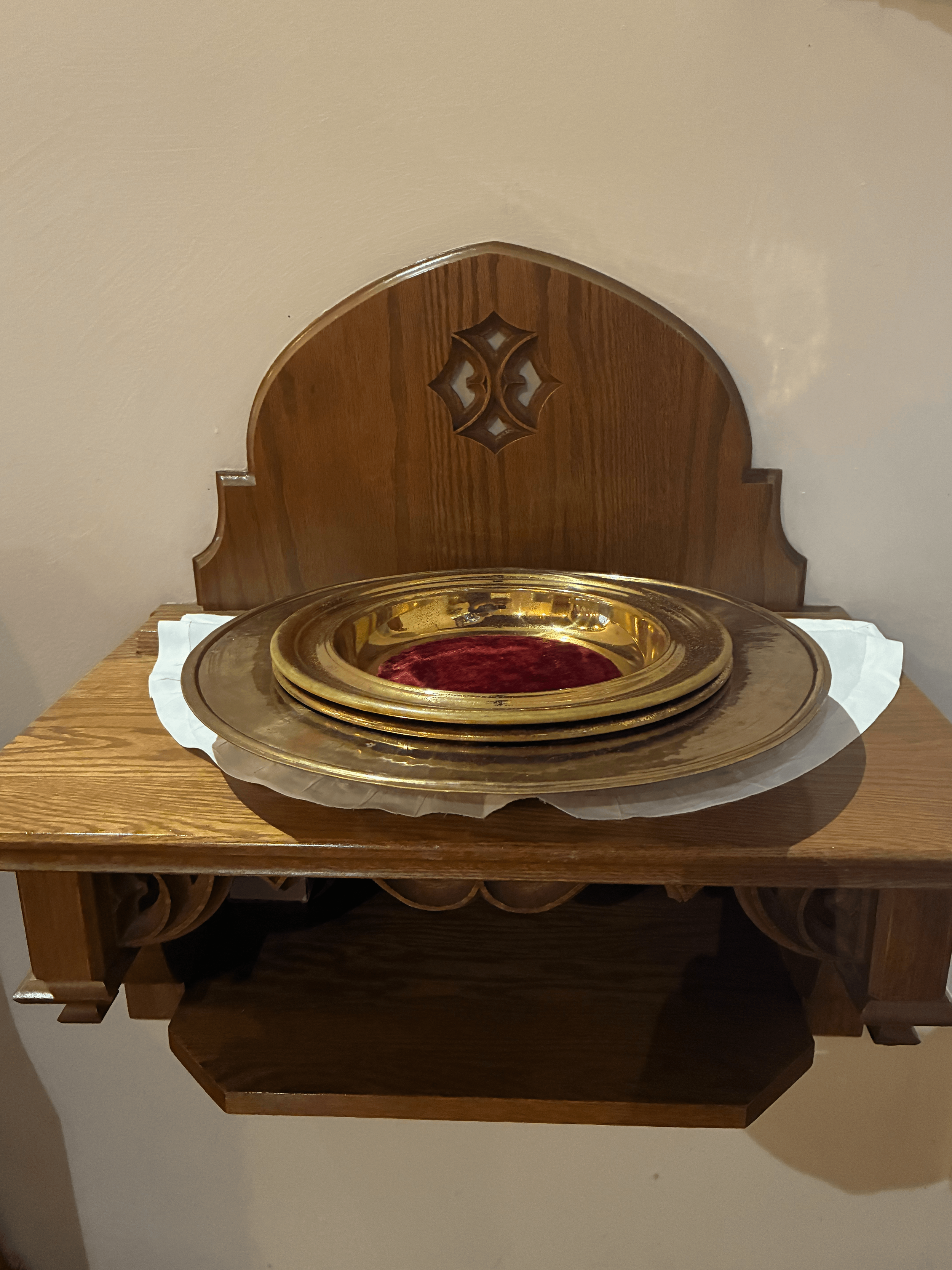 A wooden table with a decorative wooden piece mounted on a wall in the background, and a shiny, gold-colored tray with a red cloth inside placed on the table, with tissue paper on the side.