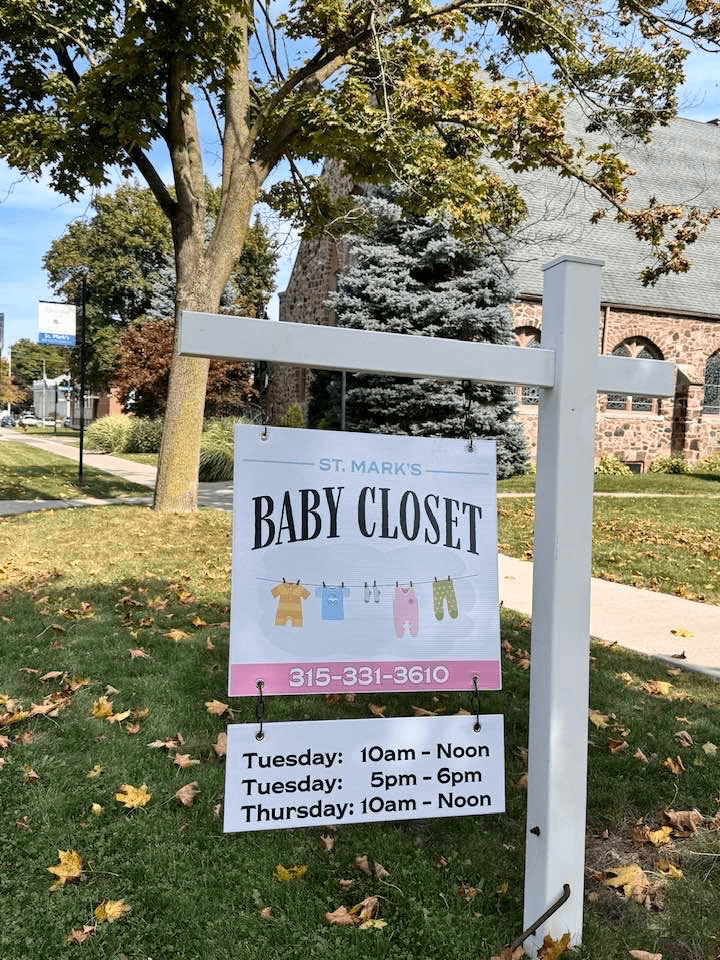 Sign for St. Mark's Baby Closet with contact number and hours of operation listed for Tuesday and Thursday, and a decorative illustration of baby clothes hanging on a line.