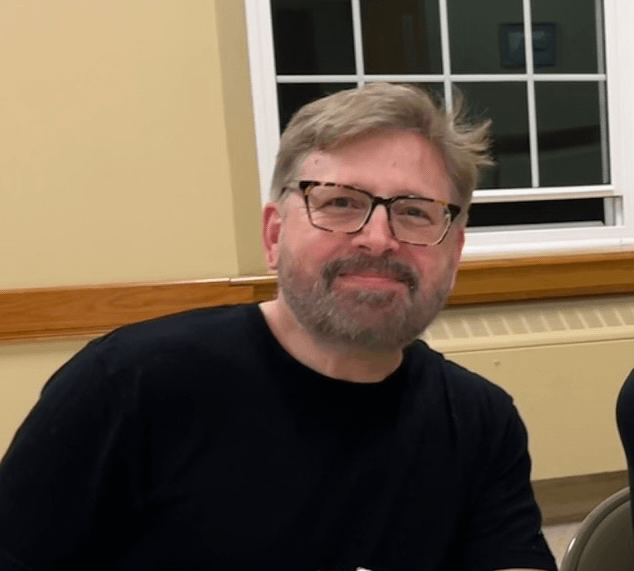 A man with glasses and a beard, smiling, wearing a black shirt, sitting indoors near a window.