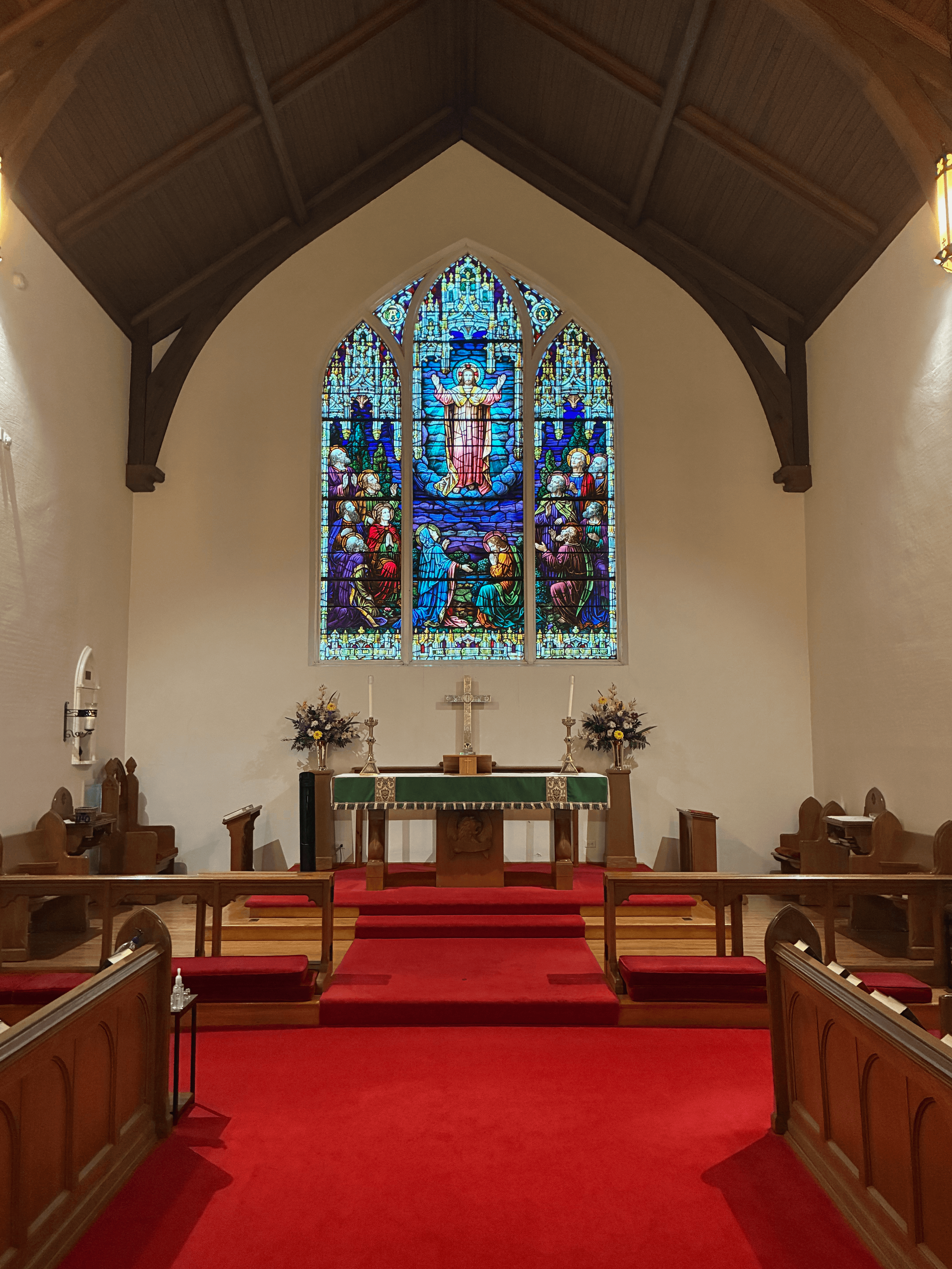Interior of a church altar with a stained glass window depicting Jesus Christ and angels above it. The altar has a green cloth, candles, and floral arrangements, with red carpeting leading up to it. Wooden pews are on either side.