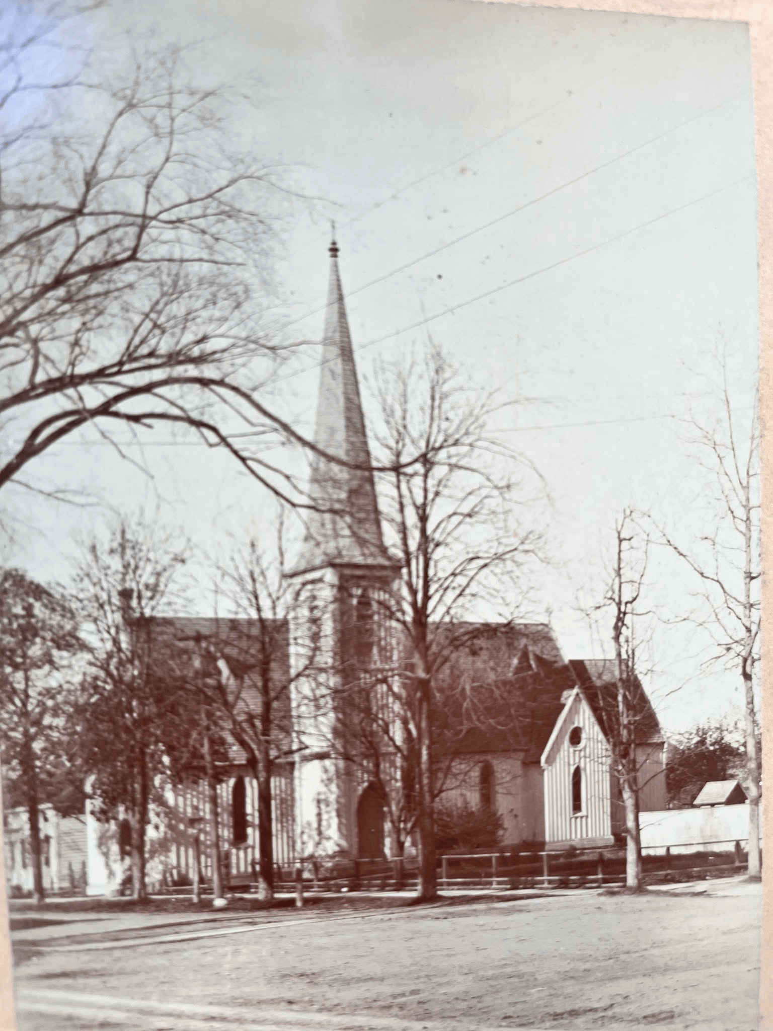 A black-and-white photograph of a church with a tall steeple, situated behind leafless trees, with a house visible in the foreground. The scene appears to be in a rural or small-town setting.