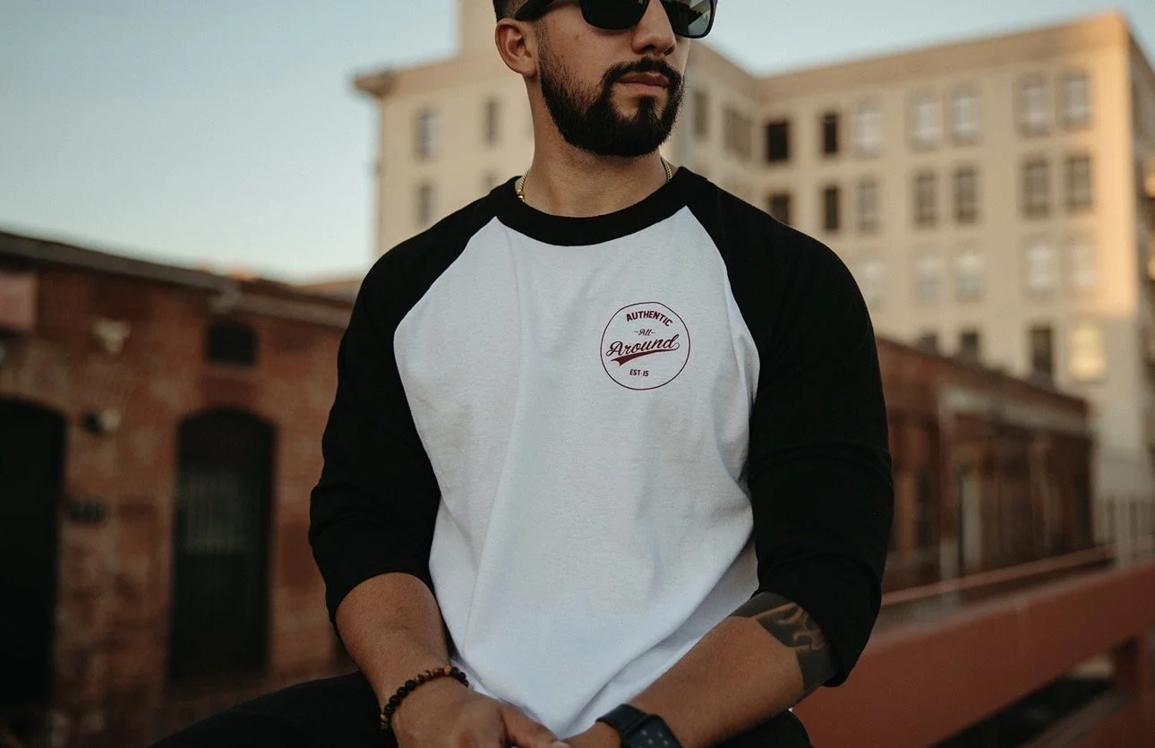 A man wearing sunglasses, a black and white baseball tee with a small patch on the chest, sitting outdoors with a brick building and a multi-story building in the background.