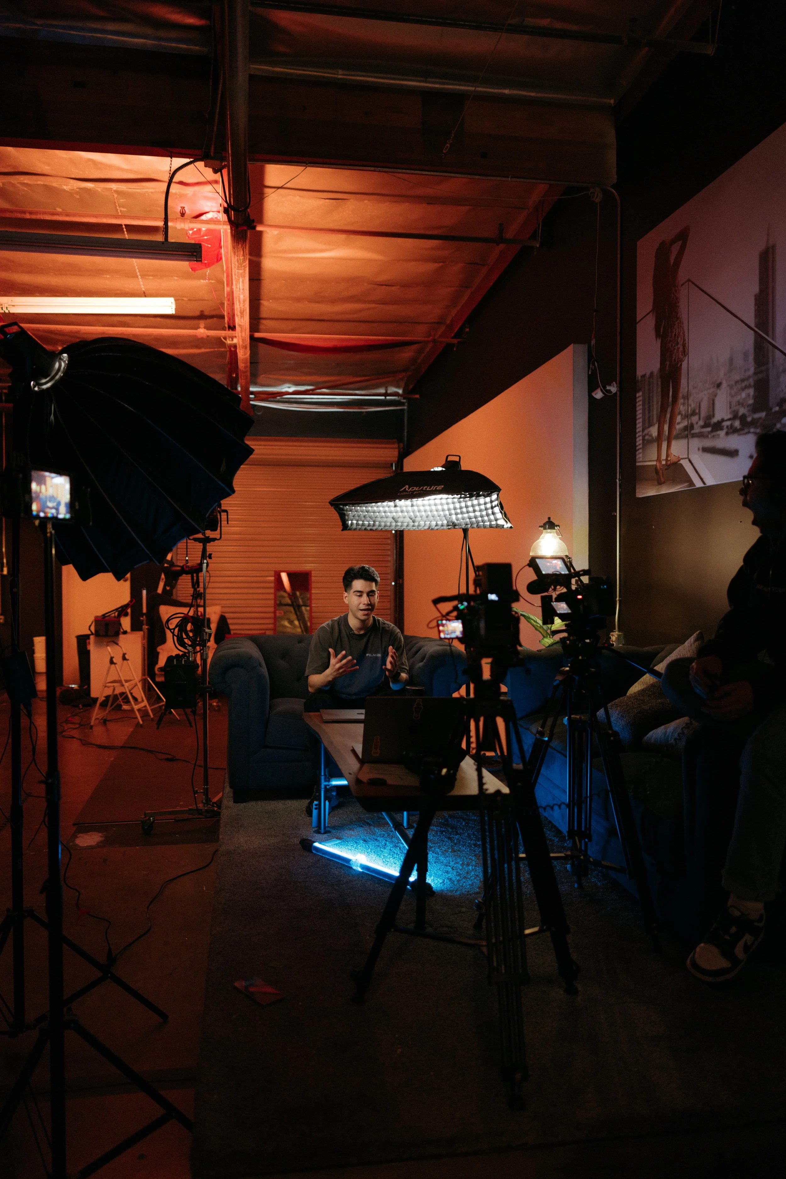 A video or photo shoot setup with professional lighting, cameras, and crew in a room with artistic decor, including a large wall portrait of a woman in a cityscape background, and a person sitting on a couch speaking or being interviewed.