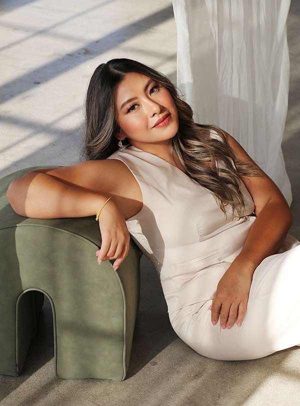 A woman with long wavy hair, wearing a sleeveless beige dress, reclining on a green upholstered armrest with sunlight streaming through a window behind her.