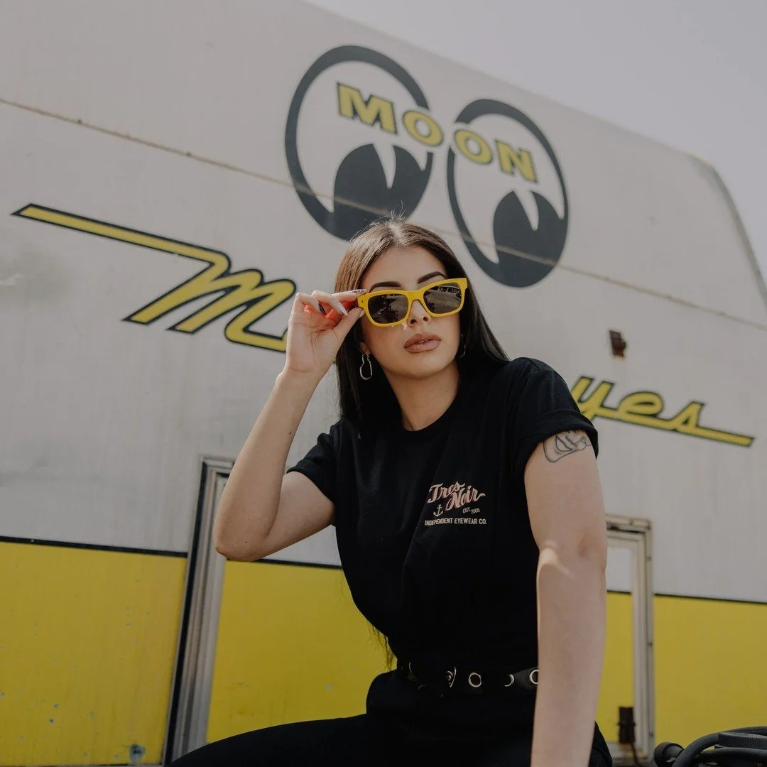 Woman with dark hair wearing yellow sunglasses, sitting outdoors in front of a large wall sign that reads 'MOOON' with a graphic of two black circles with a yellow outline at the top and stylized yellow text
