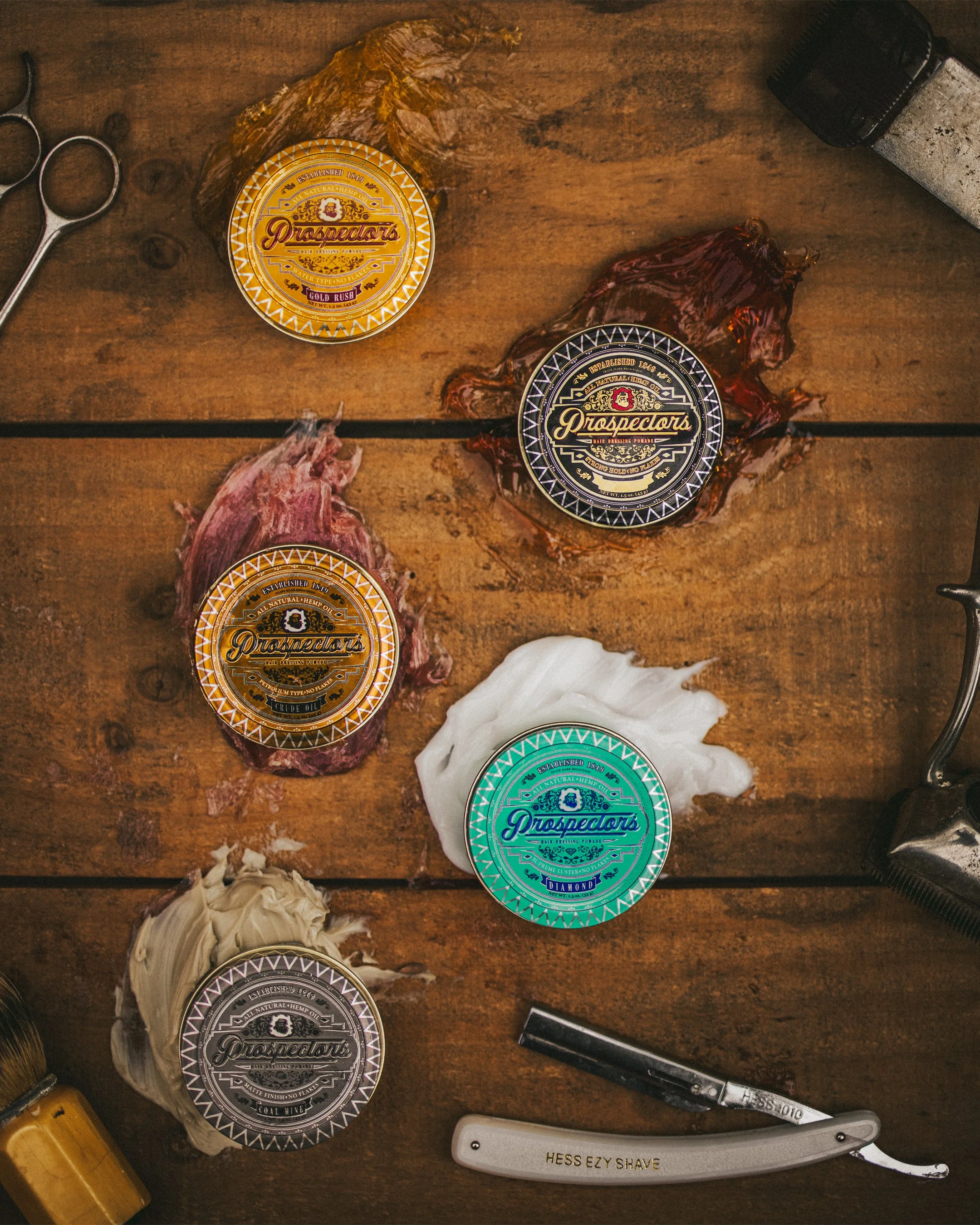 Collection of hair styling products and tools on a wooden surface, including jars of Prospector's hair dressing pomade in different scents, a shaving razor labeled Hess E Z Y Shave, scissors, and some hair or grooming wax.