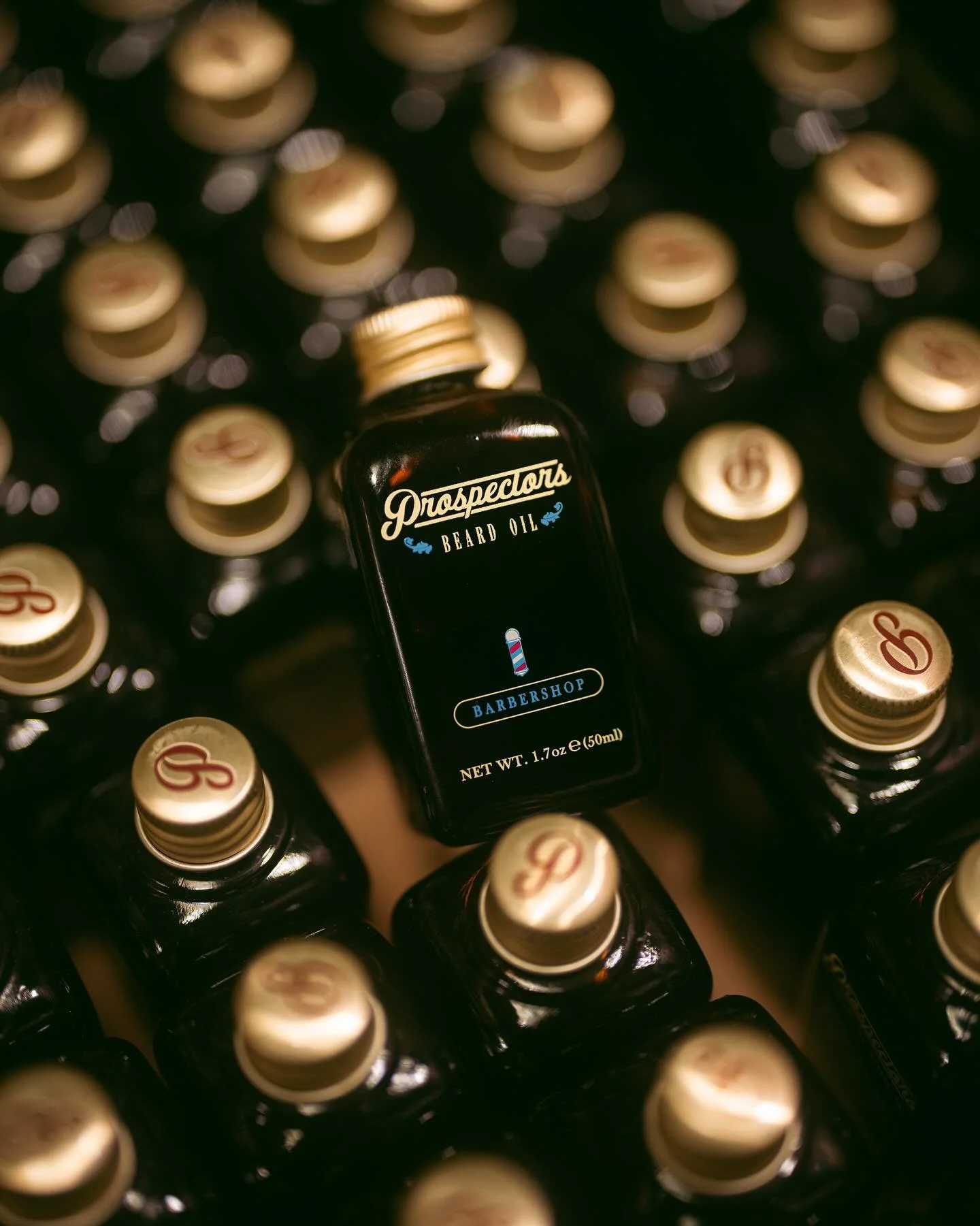 A bottle of Prospector beard oil placed among multiple bottles of G&B brand dark-colored liquid products with beige caps arranged in a grid pattern.