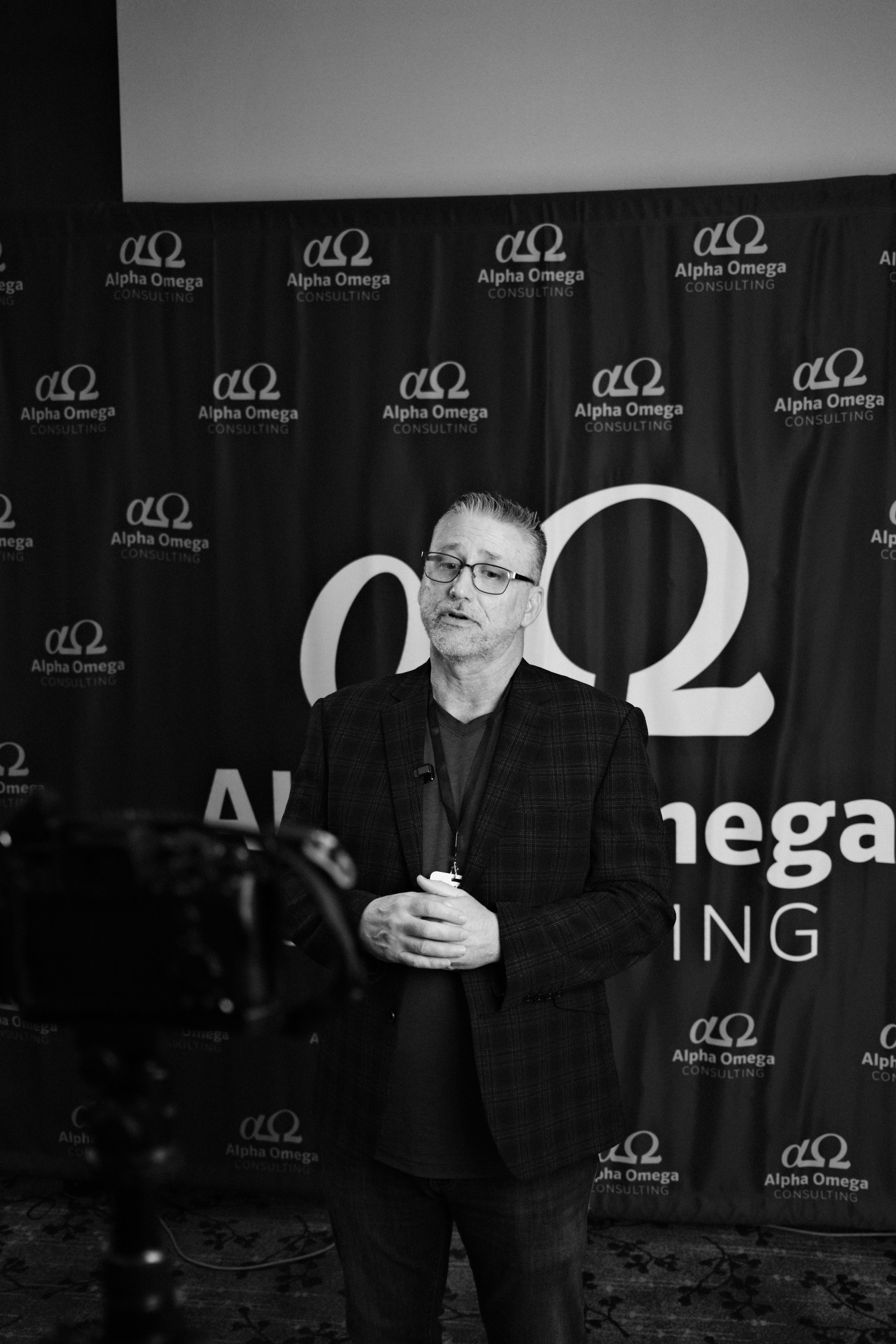 A man with glasses, wearing a checkered blazer and dark shirt, standing in front of a backdrop with the logo and name of Alpha Omega Consulting, during a presentation or interview setting.