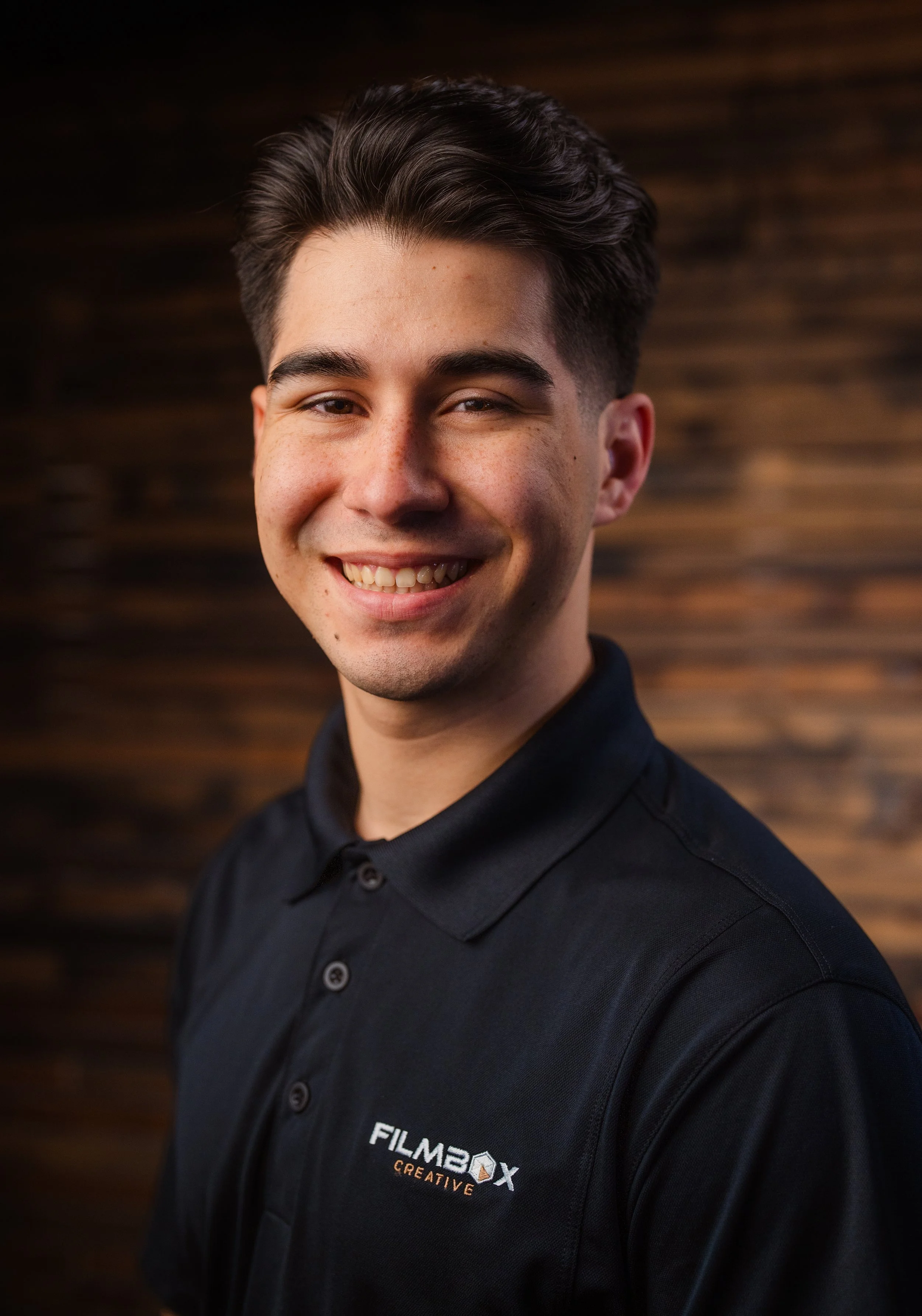 Close-up of a young man with dark hair and fair skin, smiling, wearing a black collared shirt with a logo that says 'FILMBOX CREATIVE,' standing against a blurred brown wooden background.
