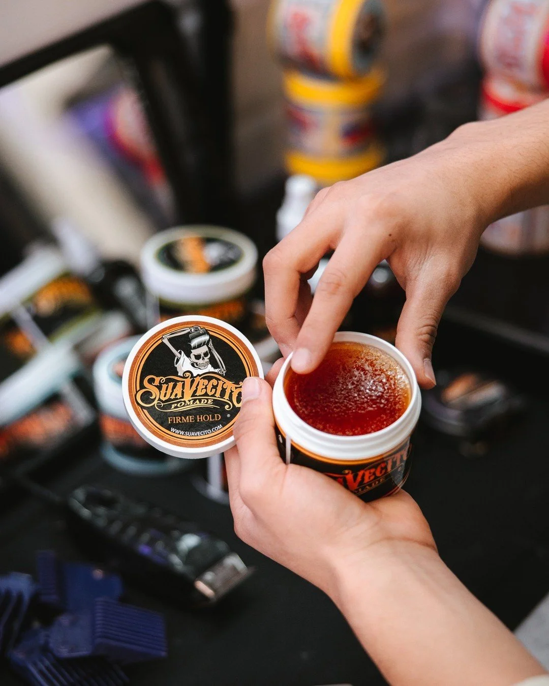A person opening a container of Suavecito Pomade, with the lid featuring a skull logo, in front of a display of other grooming products.