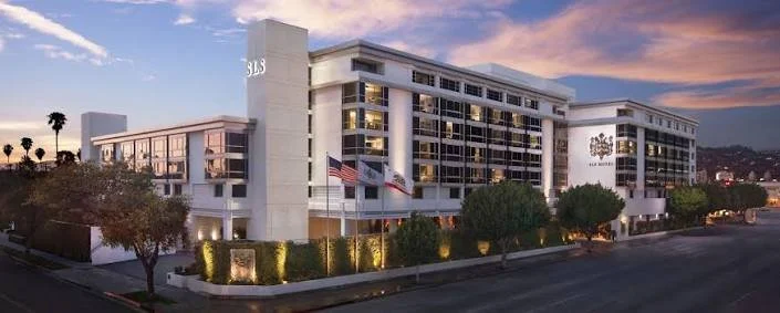 Exterior view of a modern white hotel building with multiple floors, balconies, flags, and the name 'SLS' on the side, during sunset.