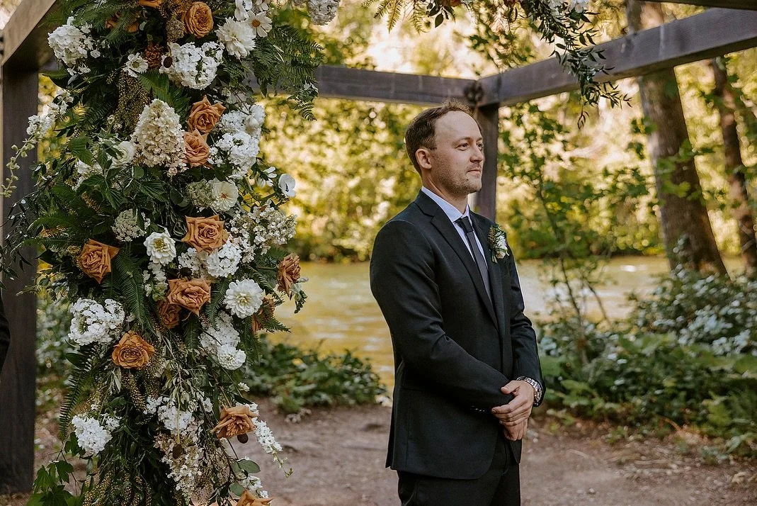A groom in a black suit standing at an outdoor wedding arch decorated with white and orange flowers near a river and surrounded by greenery.