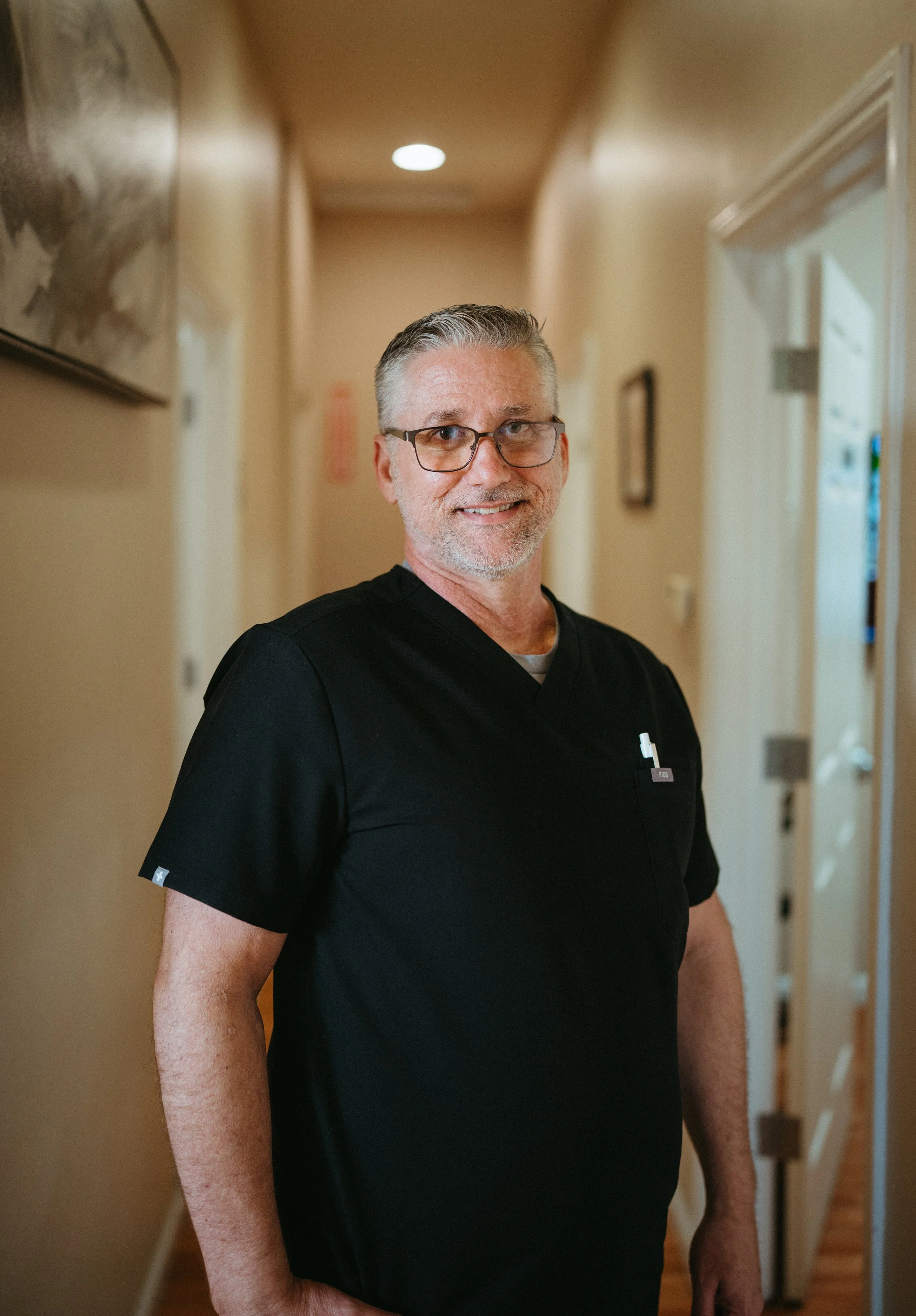 A middle-aged man with glasses, short gray hair, and a beard, wearing a black medical uniform, standing in a hallway and smiling at the camera.