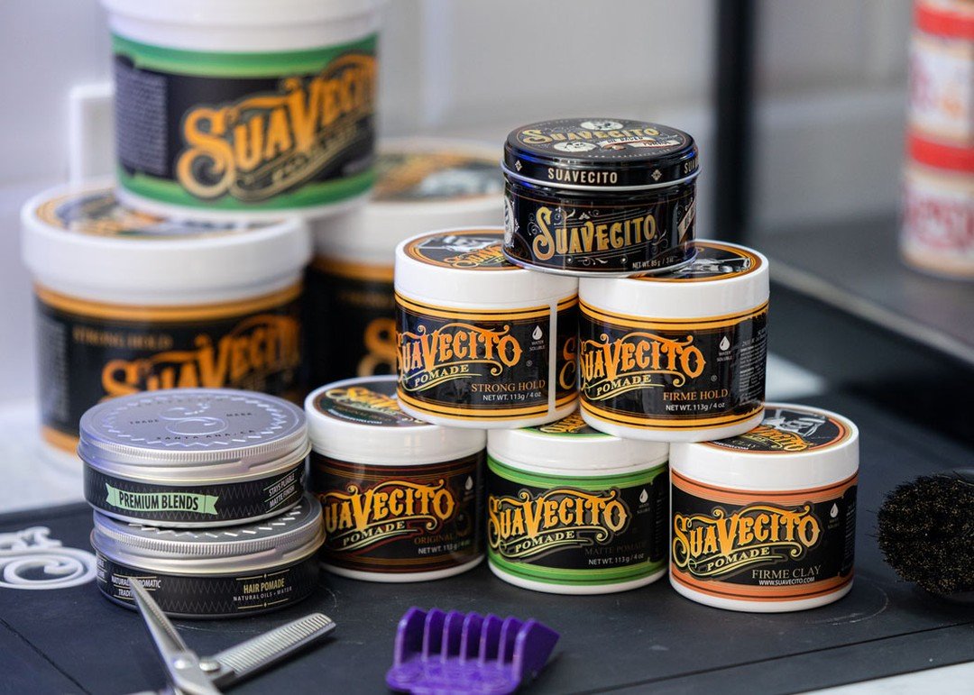 Several containers of Suavecito pomade stacked on a black surface, with some labeled as premium blend, original, matte, and firm clay, along with a purple hair comb, a pair of hair scissors, and a black scrubbing pad.