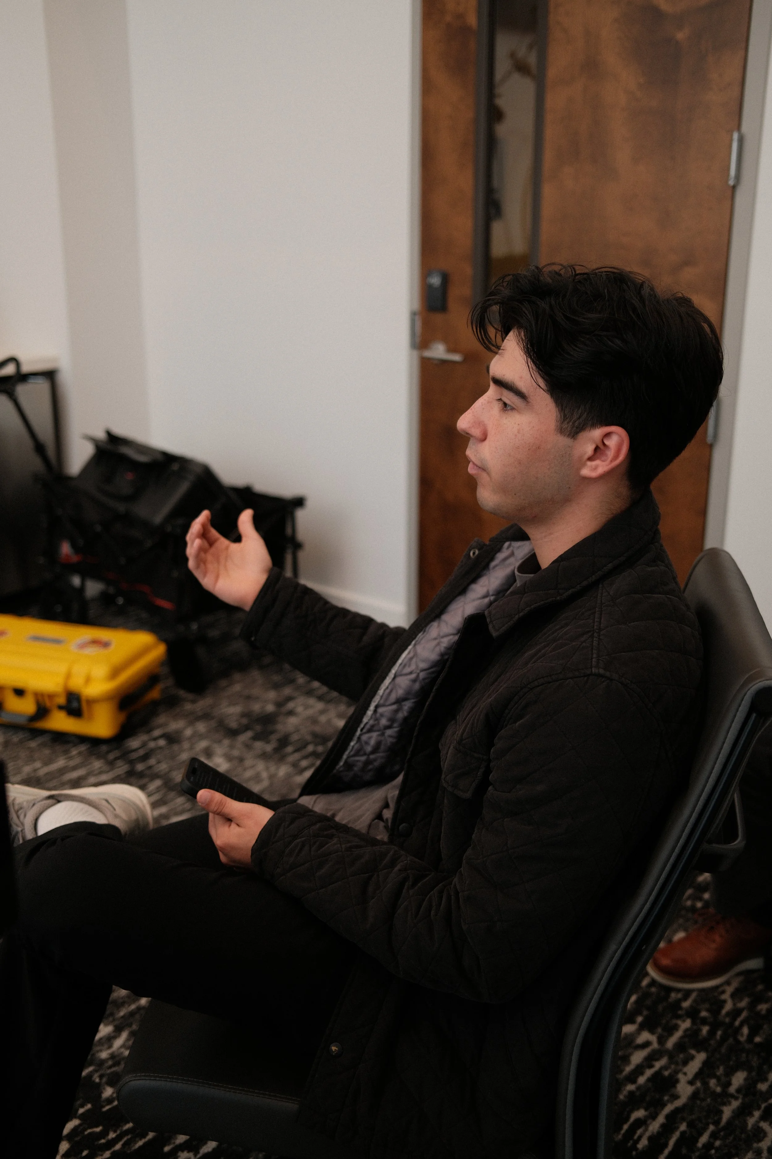 A young man with dark hair and light skin, sitting in a chair indoors, holding a remote control in his right hand, with a yellow toolbox on the table nearby. Behind him is a closed wooden door and an open closet with hanging clothes. The floor is covered with a black-and-white patterned carpet.