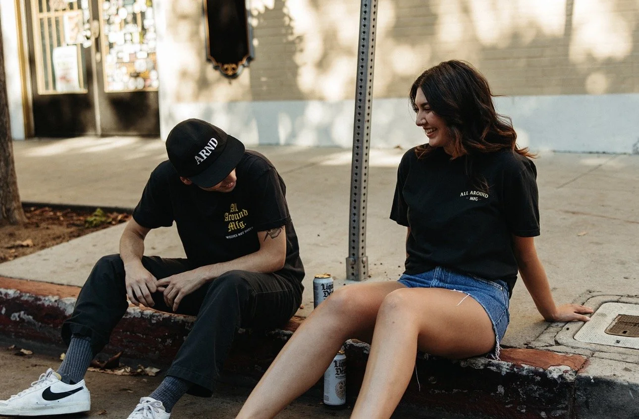 A young man and woman sitting on a curb outside, laughing and talking. The man is wearing a black cap, black t-shirt, black pants, white Nike sneakers, and has tattoos. The woman is wearing a black t-shirt and denim shorts, with her legs crossed, and has a can of beer between her legs. They appear to be enjoying each other's company on a sunny day.