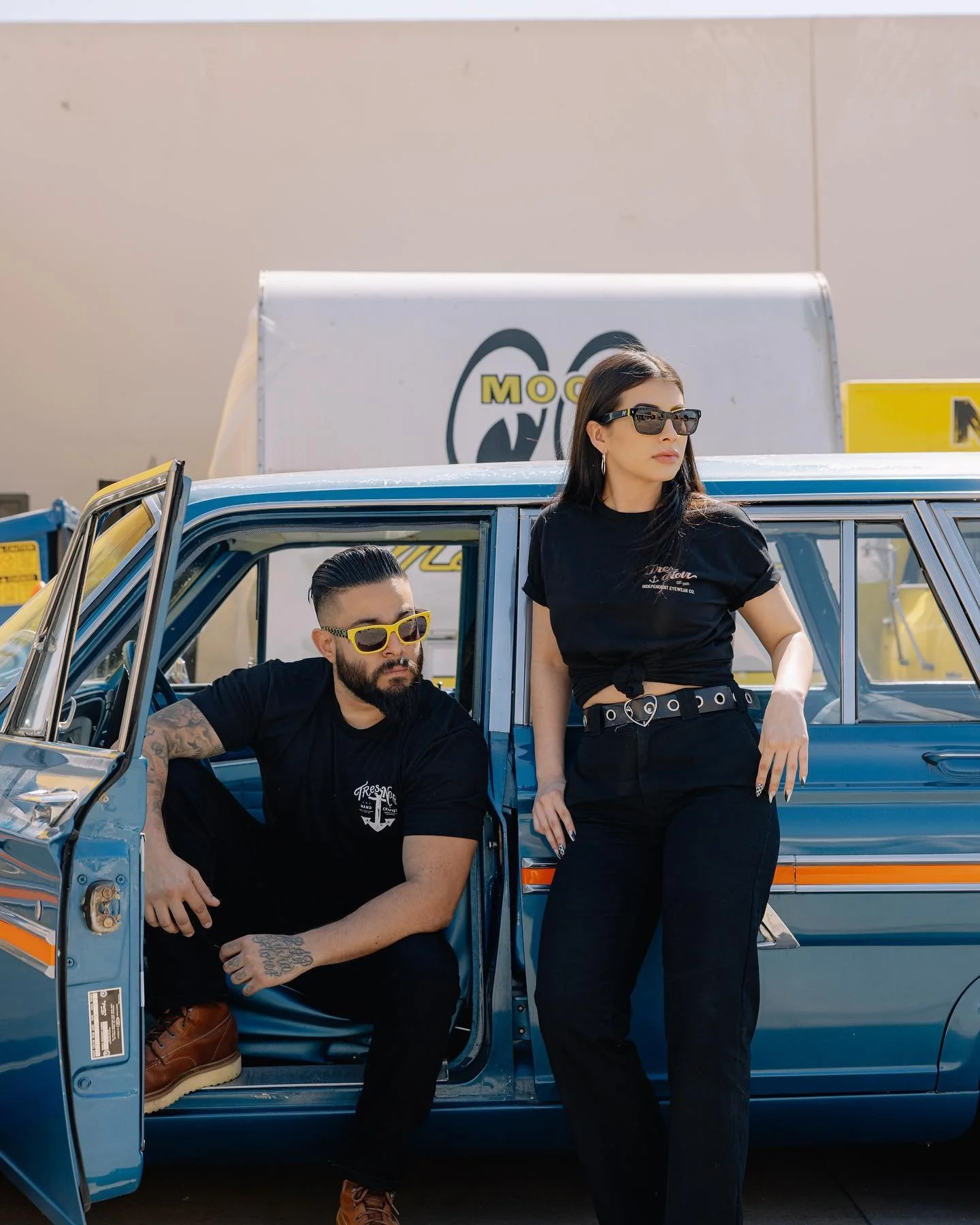 A man with a beard and tattoos wearing yellow sunglasses and a woman with dark hair and sunglasses leaning on a vintage blue car. The man is seated with his arm resting on the car door, and the woman is standing beside him with one hand on her hip. They are both dressed in black, in an outdoor setting with a large sign in the background.