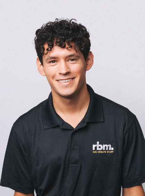 A young man with curly brown hair smiling, wearing a black polo shirt with a logo that reads 'rbm. WE CREATE STUFF' on a plain light gray background.