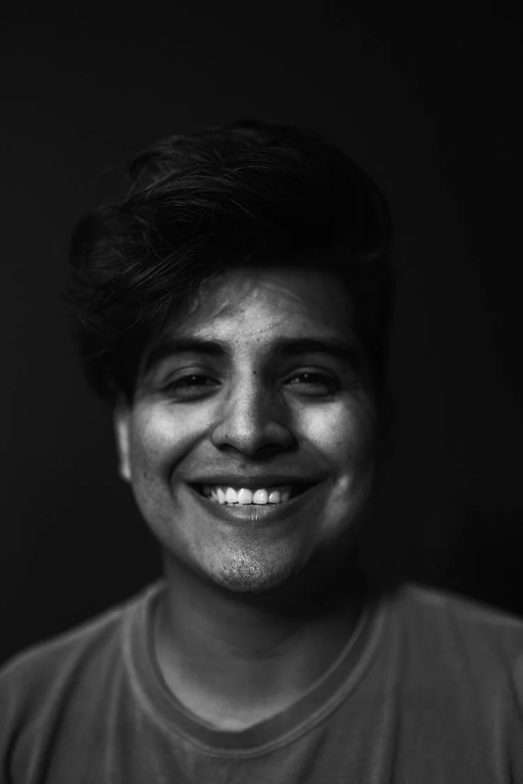 Black and white portrait of a smiling young man with short, tousled hair, wearing a casual t-shirt against a plain dark background.