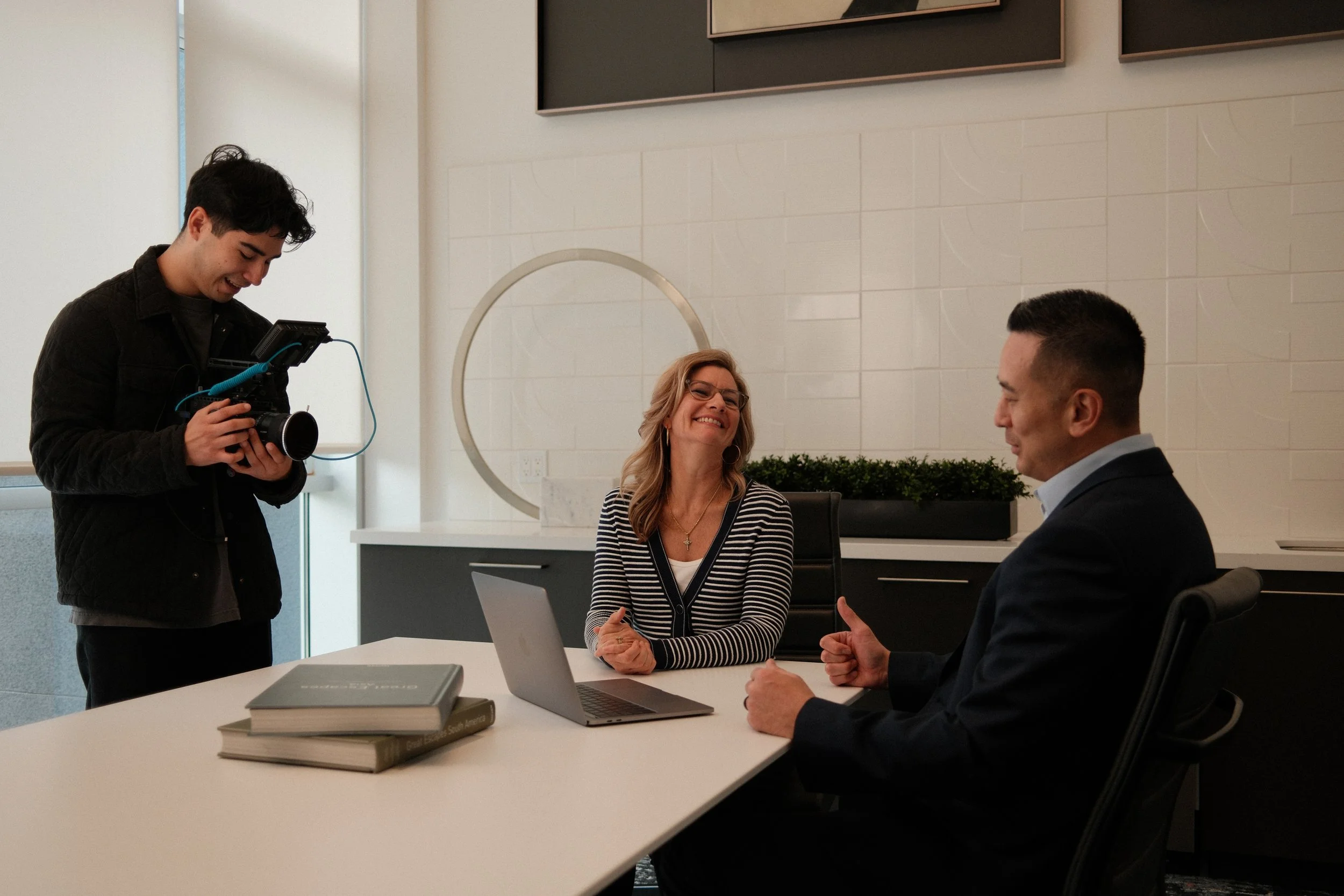 A woman with glasses and a striped blazer smiling at a man wearing a suit, sitting at a table in a modern office or conference room, while a young man with dark hair holds a camera and records the scene.