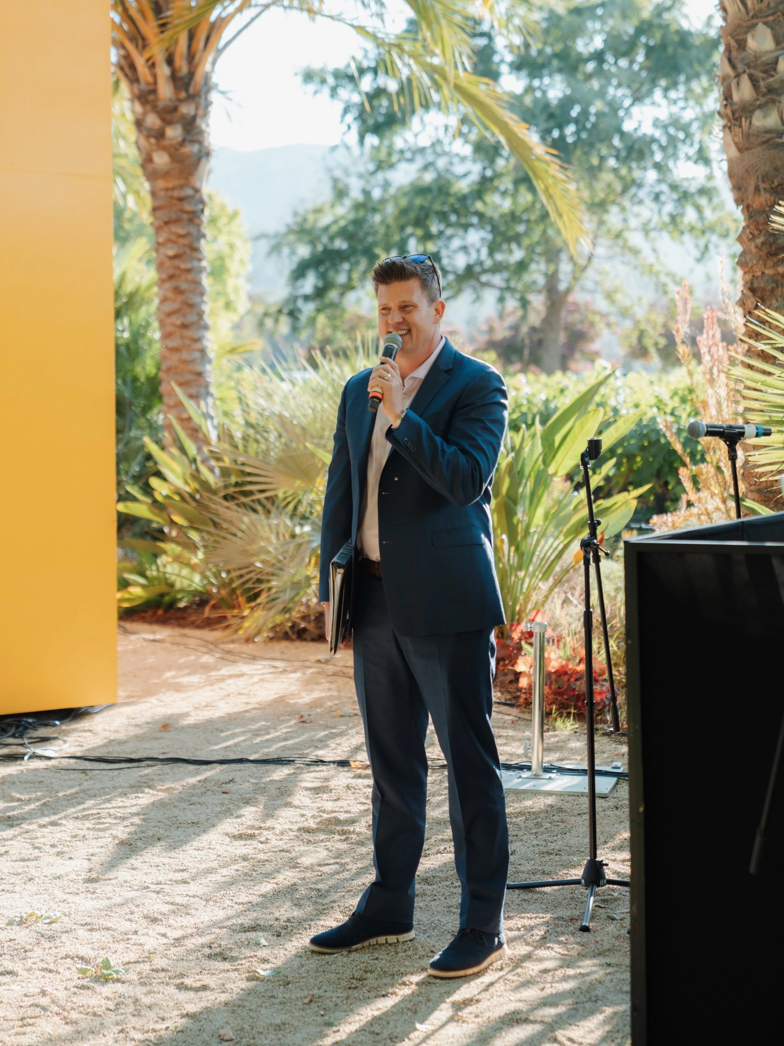 A man in a suit holding a microphone and smiling, speaking outdoors under palm trees, with a microphone stand nearby.