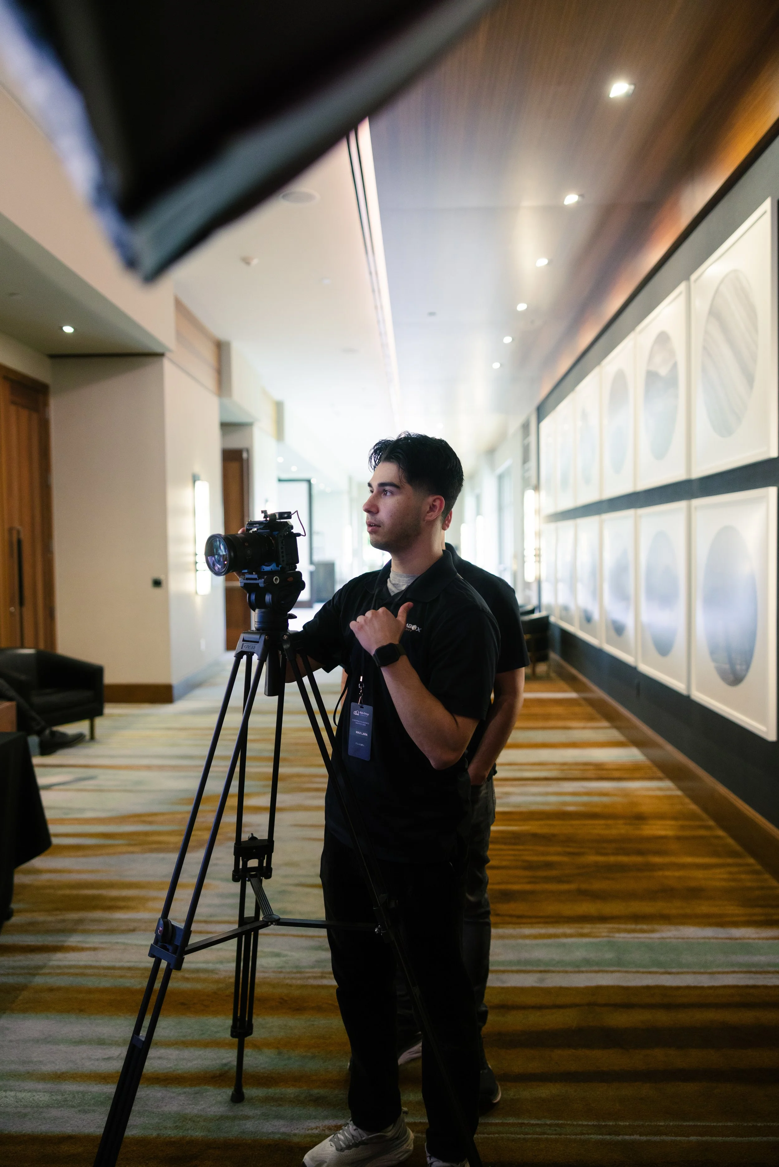 A young man in black clothing and sneakers adjusting a camera on a tripod in a conference or convention center hallway with patterned carpet and framed artwork on the wall.