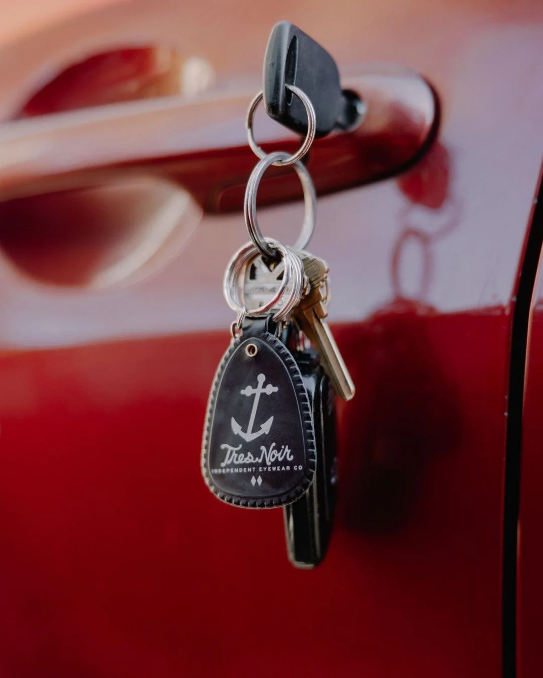 A set of keys hanging from a car door handle, which has a black keychain labeled 'Tres Noir Independent Eyewear Co' with an anchor design, on a red car door.
