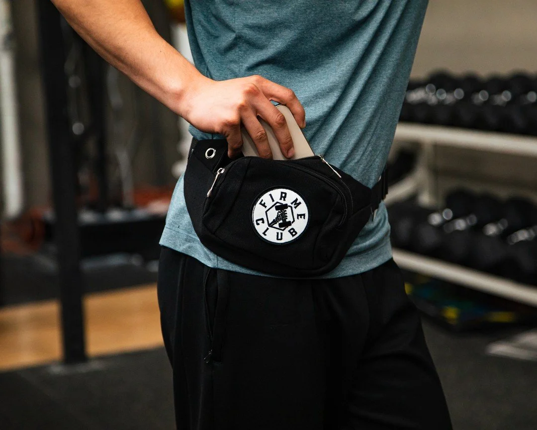 A person in workout clothes holds a small black bag with a circular Patch reading 'FIRM CLUB' in a gym with weights and exercise equipment in the background.