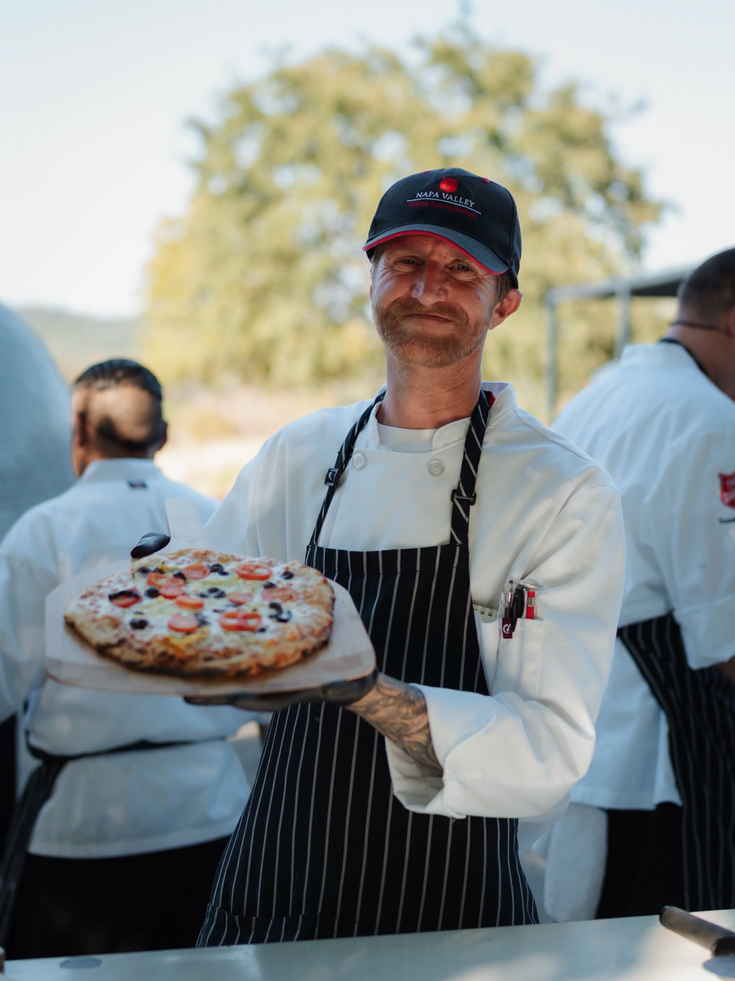 A chef with a beard and a hat labeled 'NAPA VALLEY Culinary Training Academy' is holding a pizza on a tray outdoors. The chef wears a white uniform and striped apron, and there are other chefs in similar uniforms in the background.