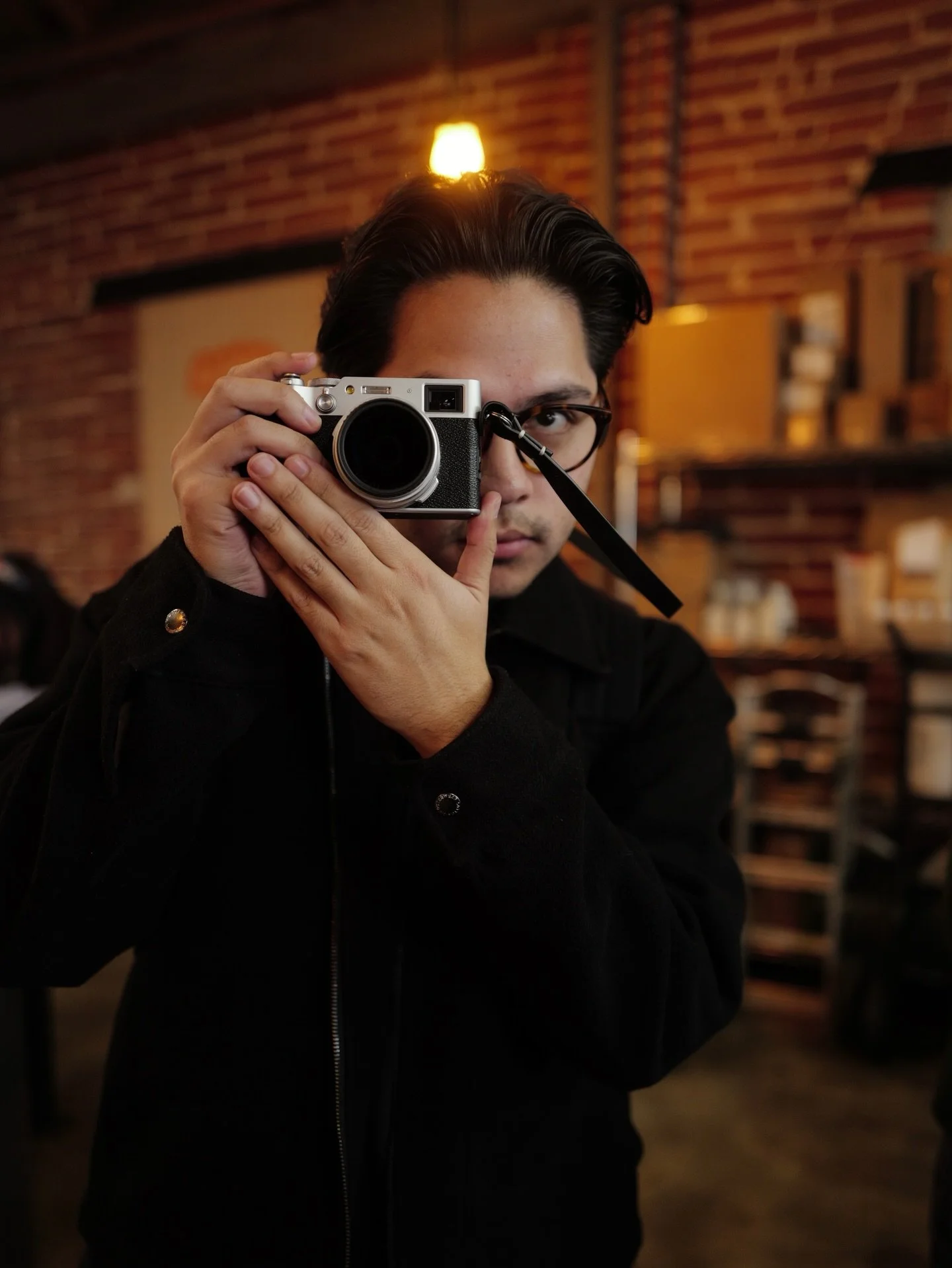 A young man with dark hair and glasses, holding a vintage-style camera up to his eye, in a dimly lit indoor setting with brick walls and warm lighting.
