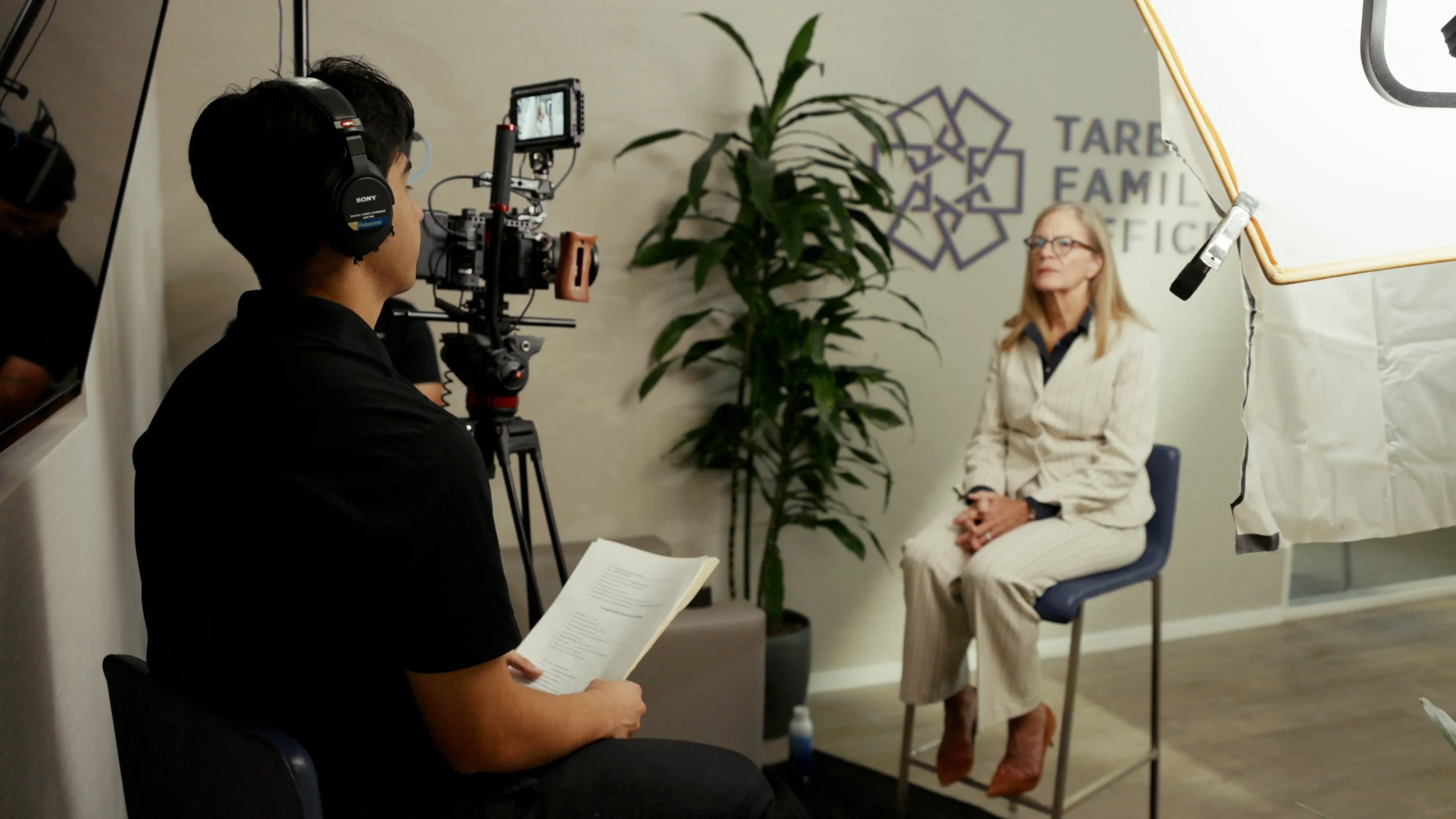 A woman with gray hair, glasses, and a striped suit sitting on a chair in a recording studio or interview setting. A camera operator is filming her with professional video equipment, and a person in the foreground is holding a script or notes. There is a sign on the wall with the logo and name of an organization and a potted plant nearby.
