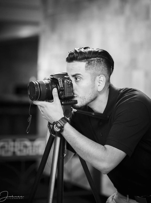 A man with styled hair and a watch taking a photograph with a camera mounted on a tripod indoors.