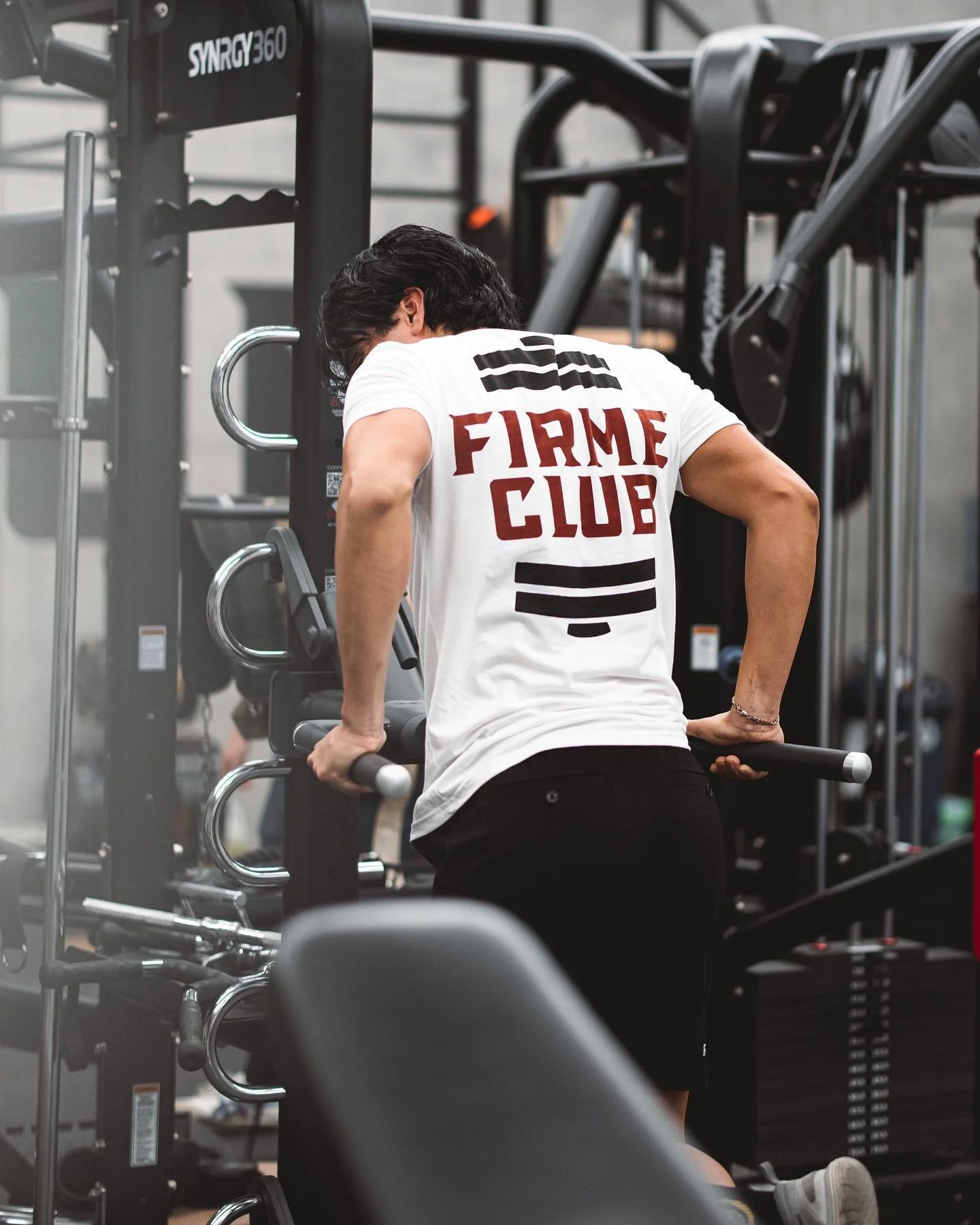 Person performing tricep dips on parallel bars at the gym, wearing a white shirt with 'FIRME CLUB' printed on the back.