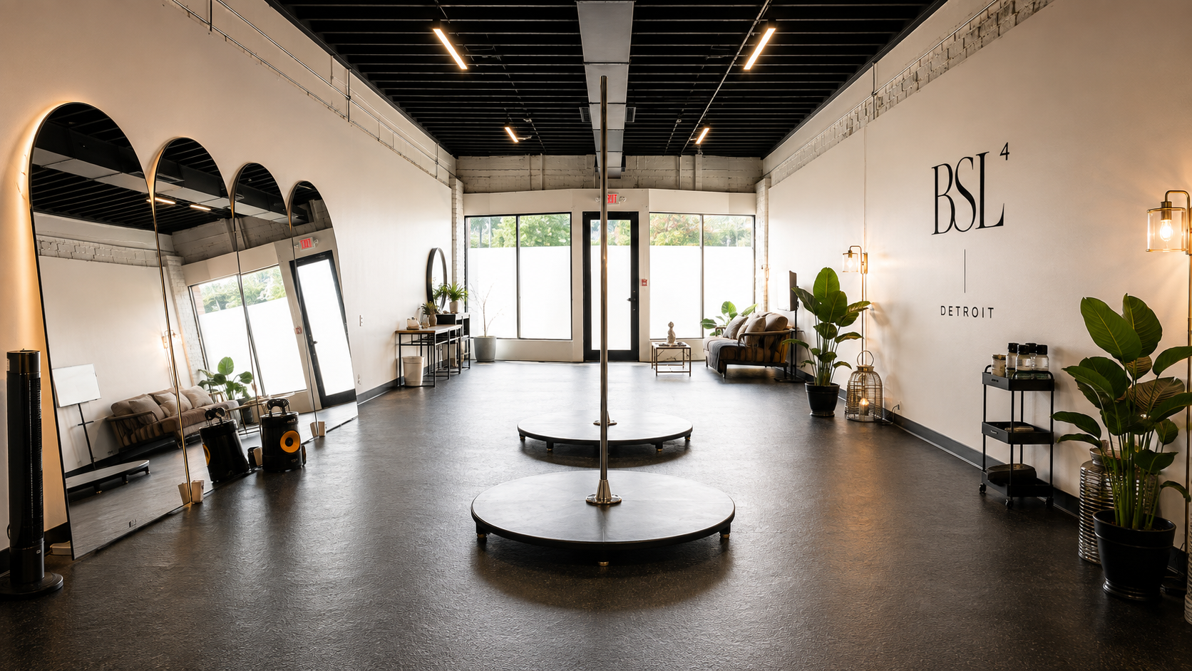 Interior of a modern, minimalist dance or fitness studio with a pole in the center, large mirrors on the left wall, and the words 'BSL 4 Detroit' on the right wall. Decor includes potted plants, seating with cushions, and warm lighting.