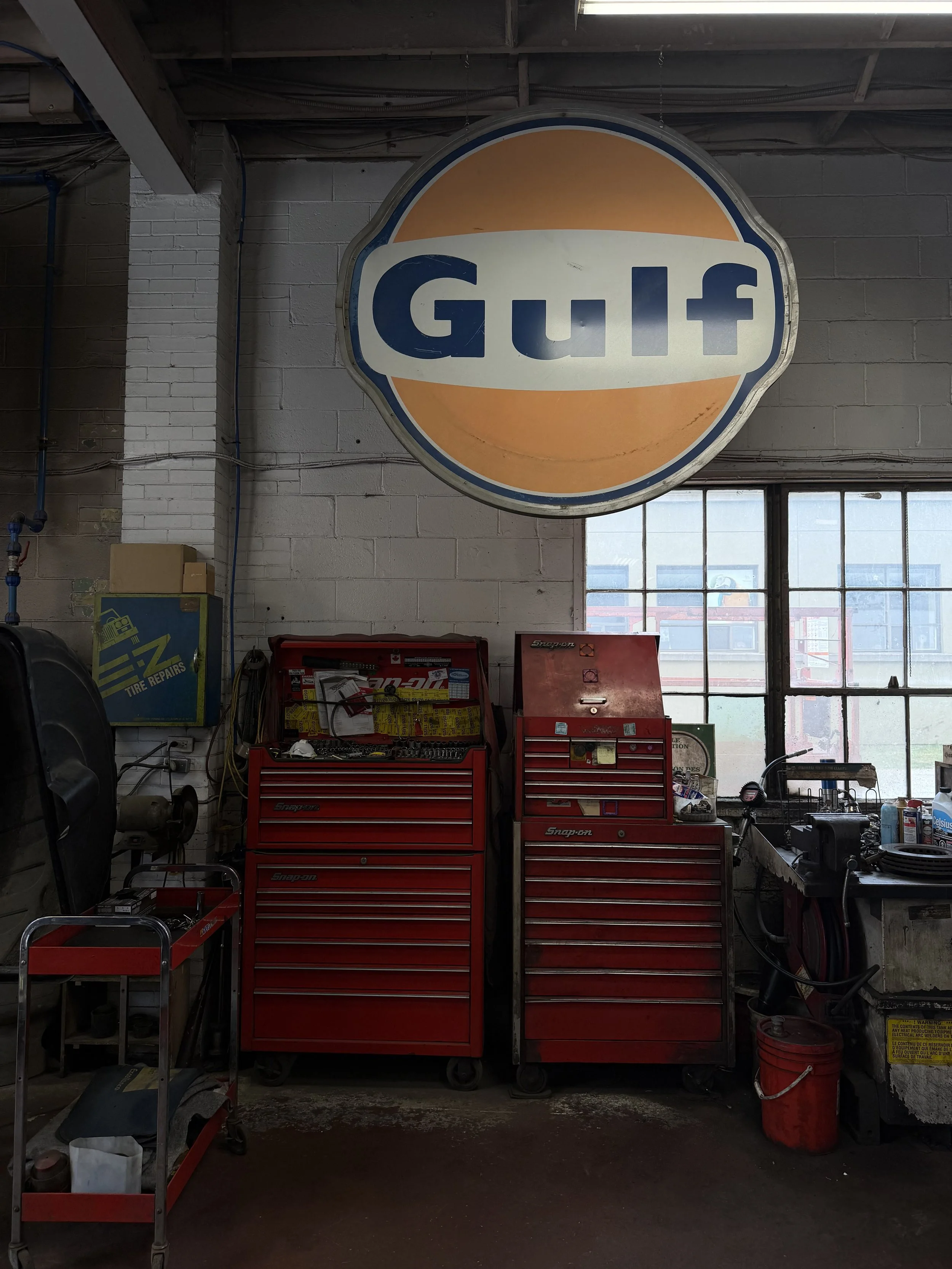 A garage workshop with a large Gulf sign hanging on the wall. Below the sign, there are two red tool chests, one with multiple drawers. The workshop contains various tools and equipment, and a window on the right side allowing natural light.