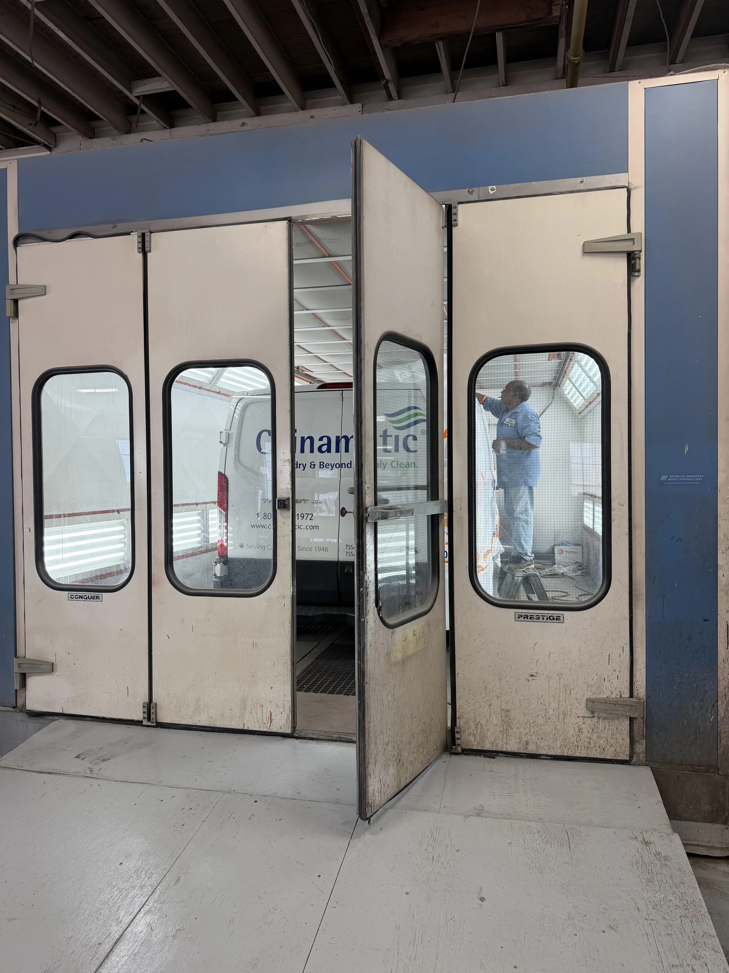 View through glass doors showing a person in a blue uniform cleaning or inspecting a ceiling with a tool, inside a commercial or industrial space with a white vehicle in the background.