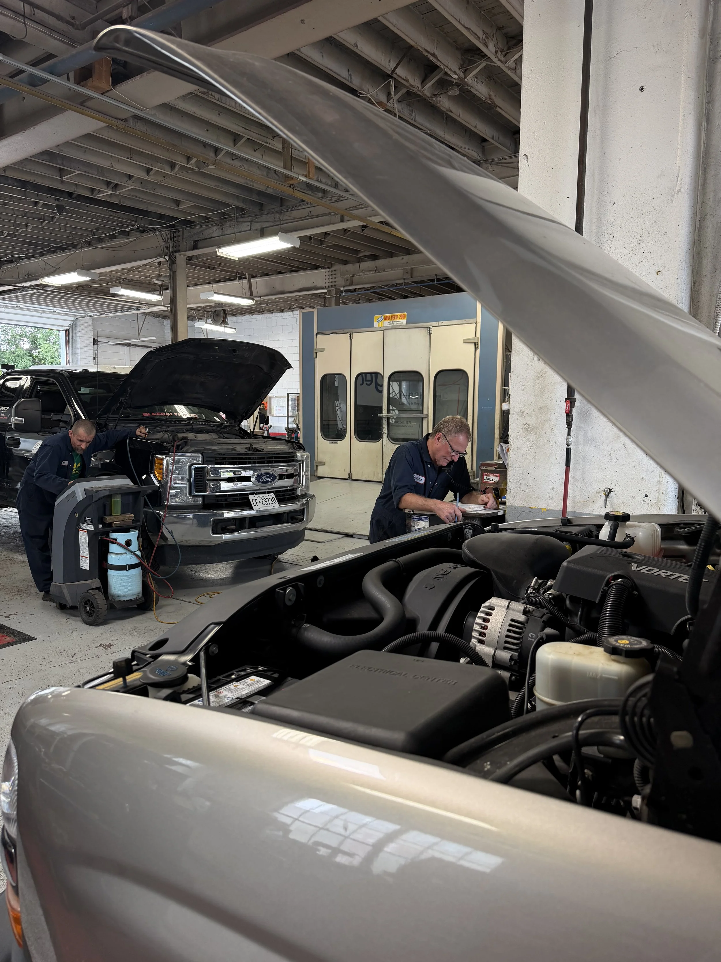Two automotive technicians working on vehicles inside a garage or workshop. One person is inspecting a black truck with its hood open, while another is writing notes or recording information near the front of a silver vehicle. The background features a large industrial air conditioning or ventilation unit, exposed ceiling beams, and bright overhead lighting.