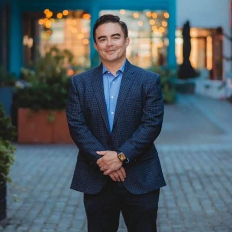 A man in a blue checkered suit stands outside on a brick-paved area, smiling at the camera with his hands clasped in front of him. The background features colorful lights, plants, and buildings, suggesting an urban or outdoor dining setting during evening.