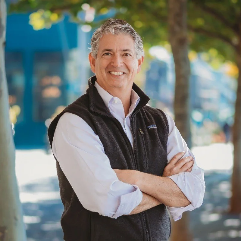 A smiling middle-aged man with curly gray hair wearing a white button-down shirt and a black vest, standing outdoors on a sidewalk with trees and a blue building in the background.
