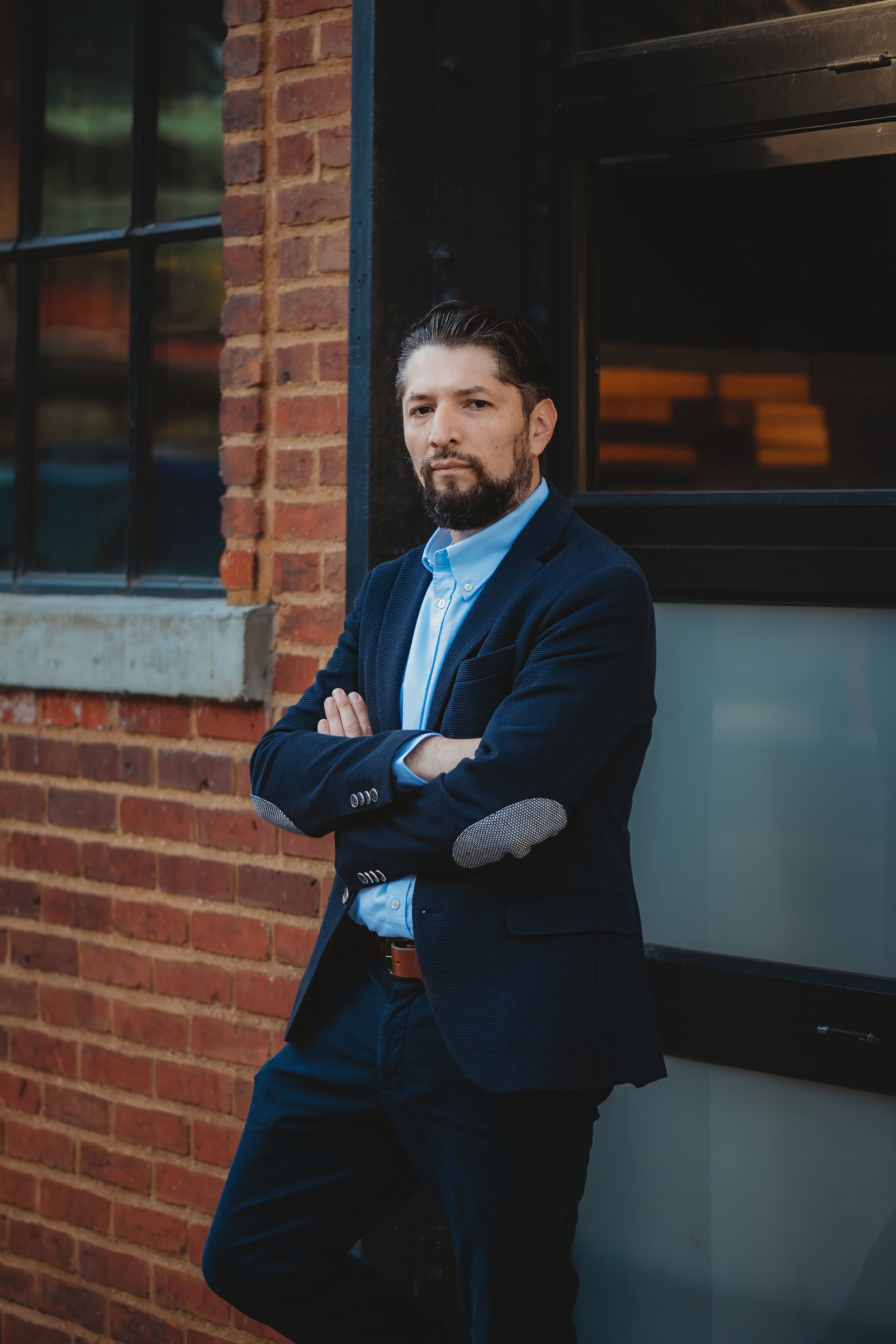 A man with a beard and slicked-back hair, wearing a dark blazer and light blue shirt, standing with arms crossed in front of a brick building with black window frames.