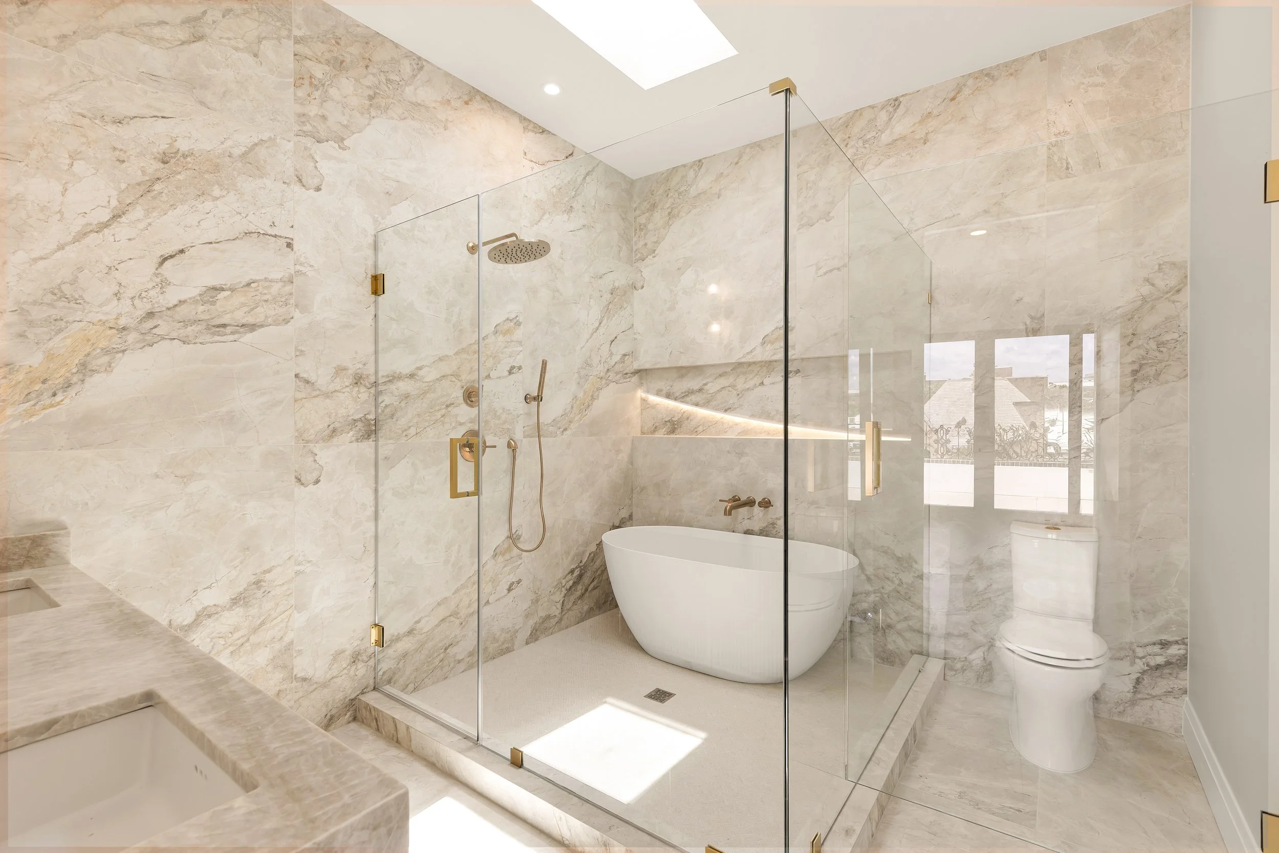 Modern bathroom featuring a glass-enclosed shower with a rainfall showerhead, a freestanding bathtub, marble walls, and large windows.