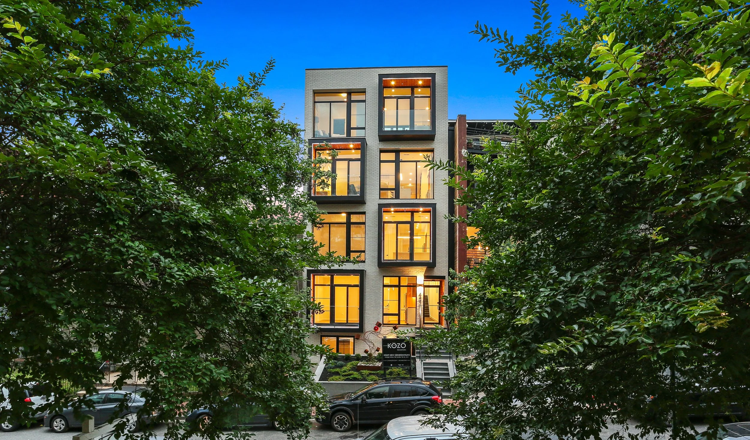 A modern multi-story building with large windows is viewed through green trees at twilight, with a parking lot and cars in front.