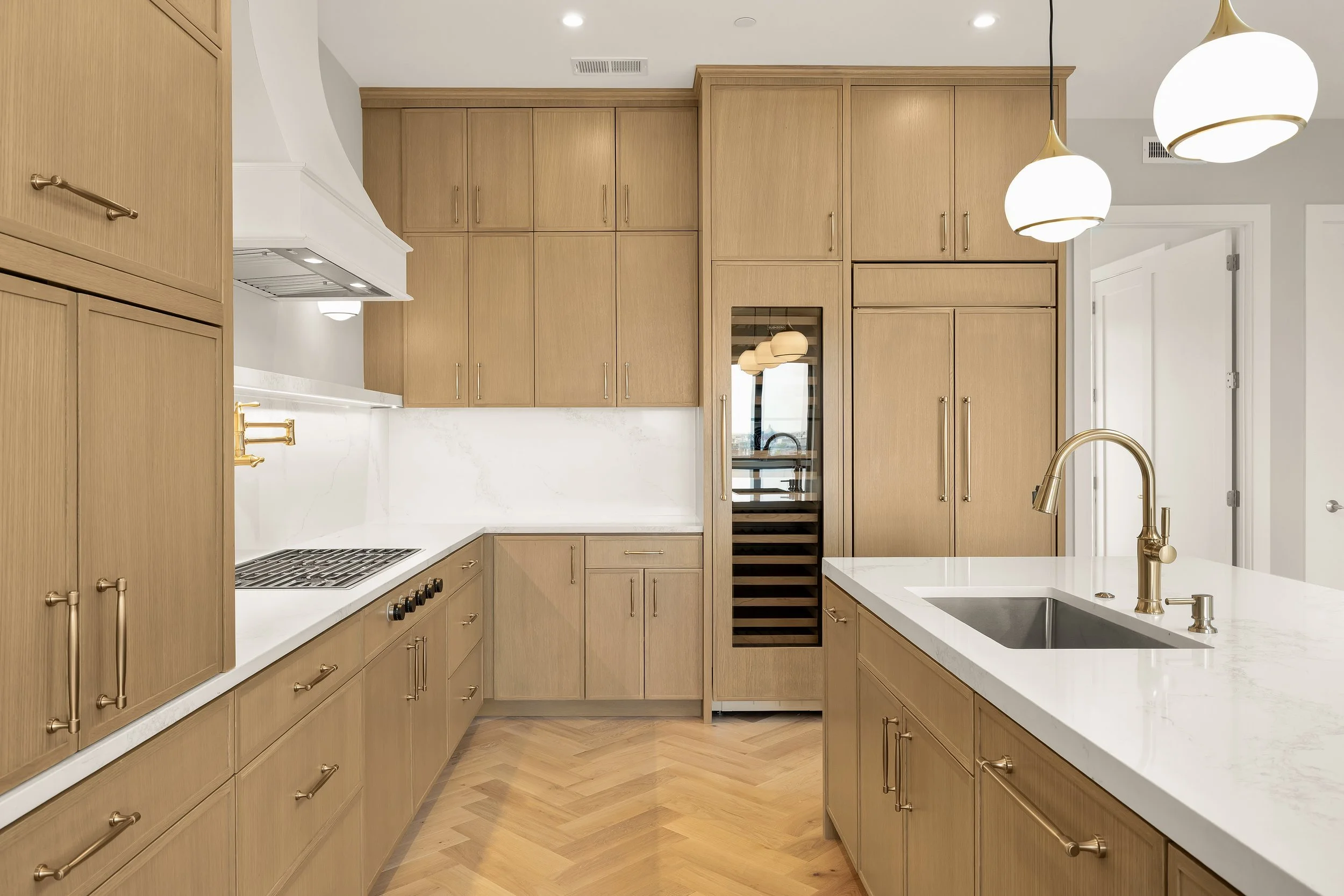 Modern kitchen with light wood cabinetry, white quartz countertops, gold hardware, built-in wine fridge, and hanging white globe pendant lights.
